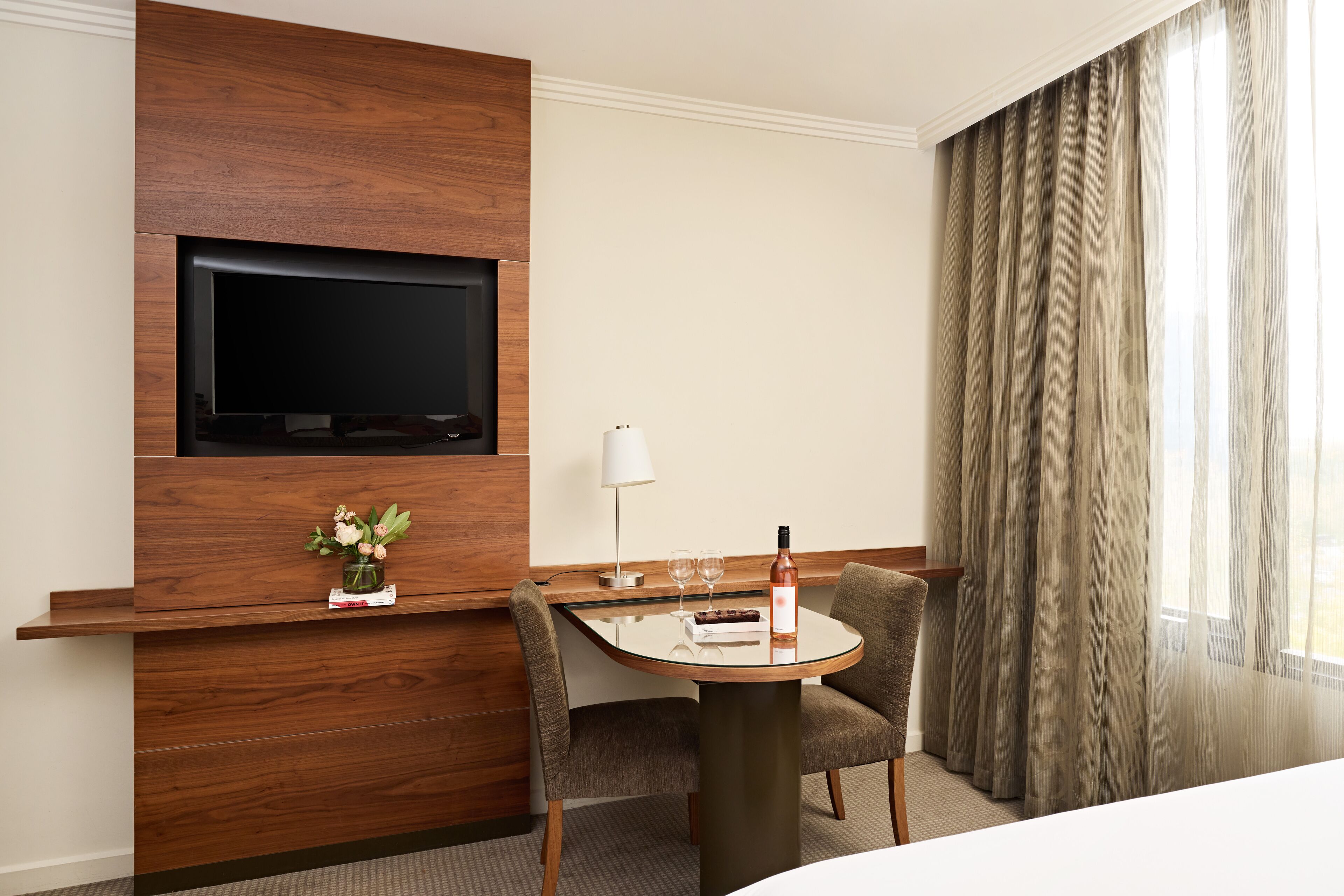 A hotel room with a flat-screen TV mounted on a wooden panel, a small table with a vase of flowers, two chairs, a bottle of wine, and a lamp.