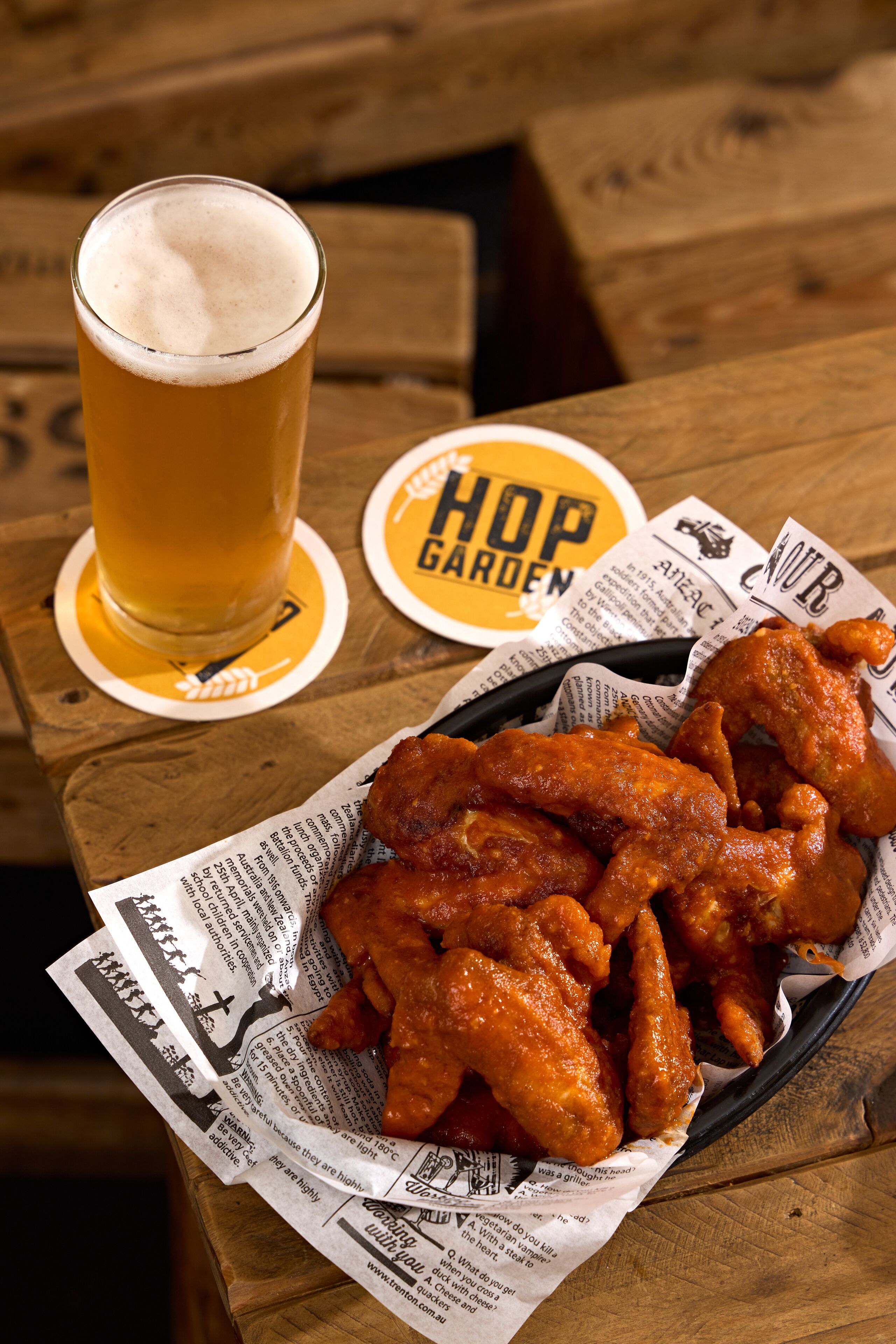 Ein Holztisch mit einem Bierglas, einer Korb gefüllt mit Chicken Wings und einem Untersetzer mit dem Schriftzug 'Hop Garden'.