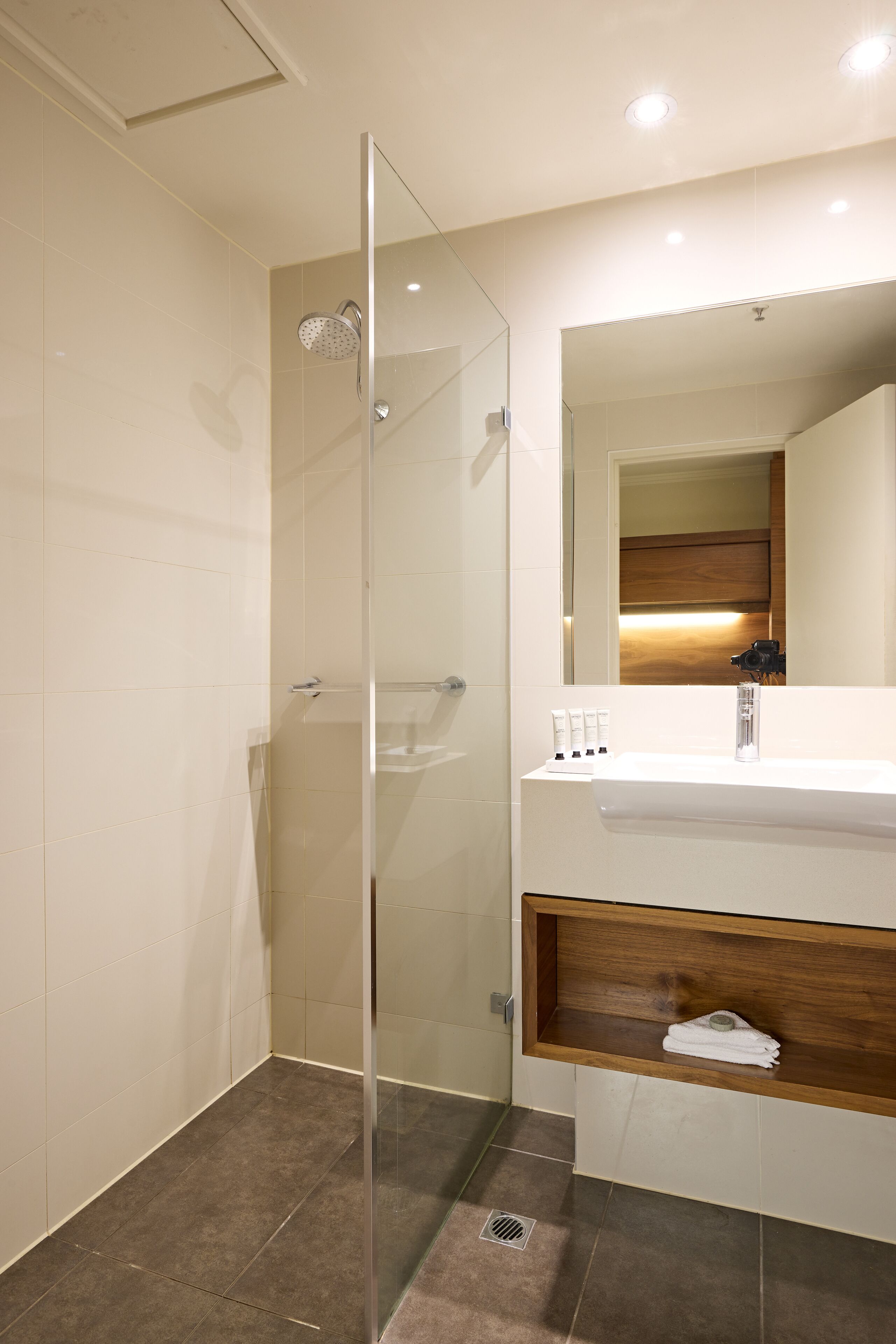 Un baño moderno con ducha envuelta en vidrio, un lavamanos blanco con estante de madera y un espejo sobre el lavamanos.