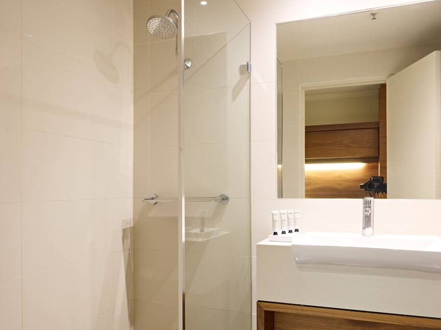 Un baño moderno con ducha envuelta en vidrio, un lavamanos blanco con estante de madera y un espejo sobre el lavamanos.