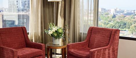 A hotel room with two red armchairs, a small round table with a vase of flowers, a floor lamp, and a view of a cityscape through large windows.