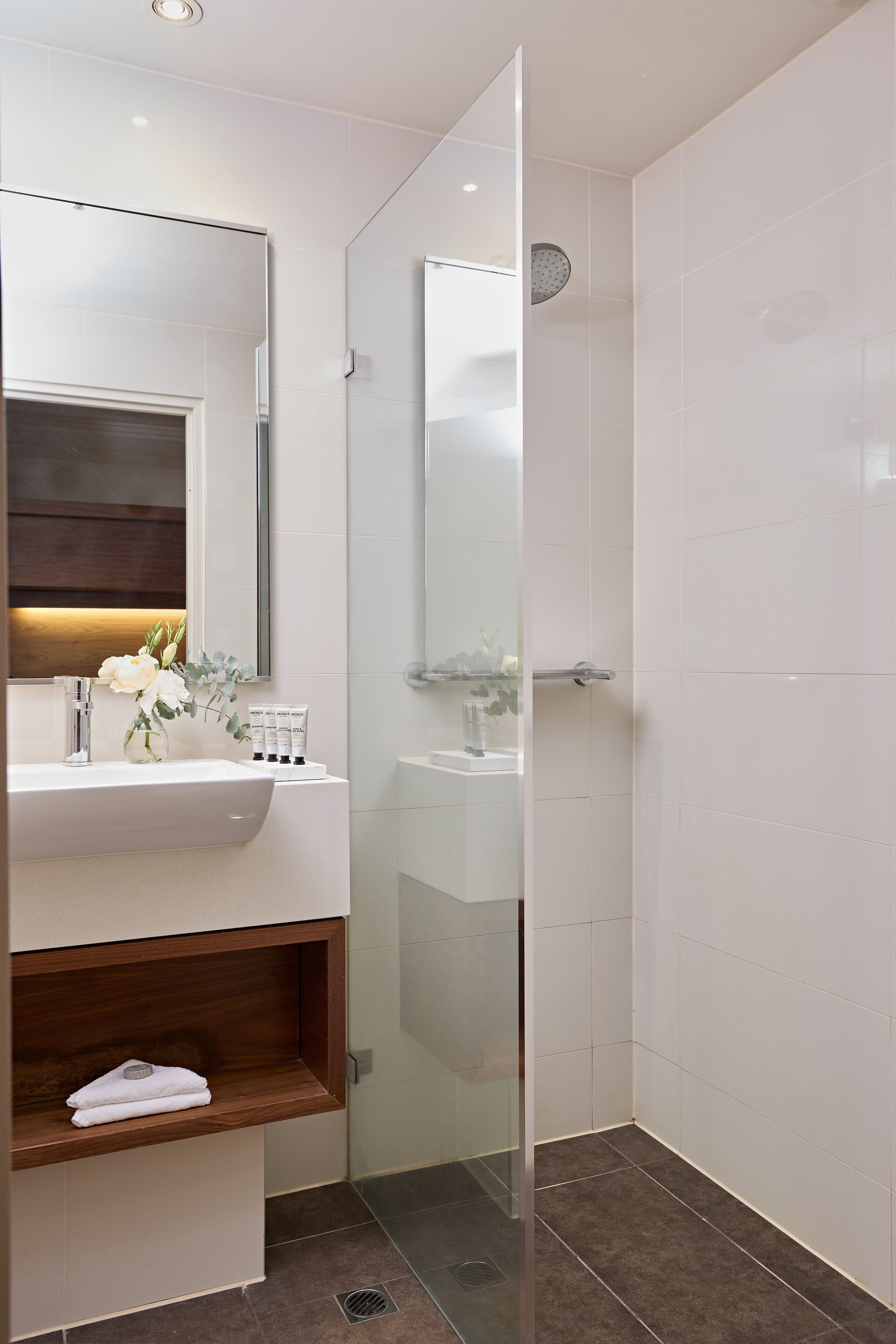 Un baño moderno con ducha de vidrio, un lavamanos blanco con espejo y un estante de madera con toallas.