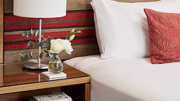 A neatly made hotel room with a bed, white pillows, a red patterned pillow, a bedside table with a lamp, a vase with flowers, and a stack of books.