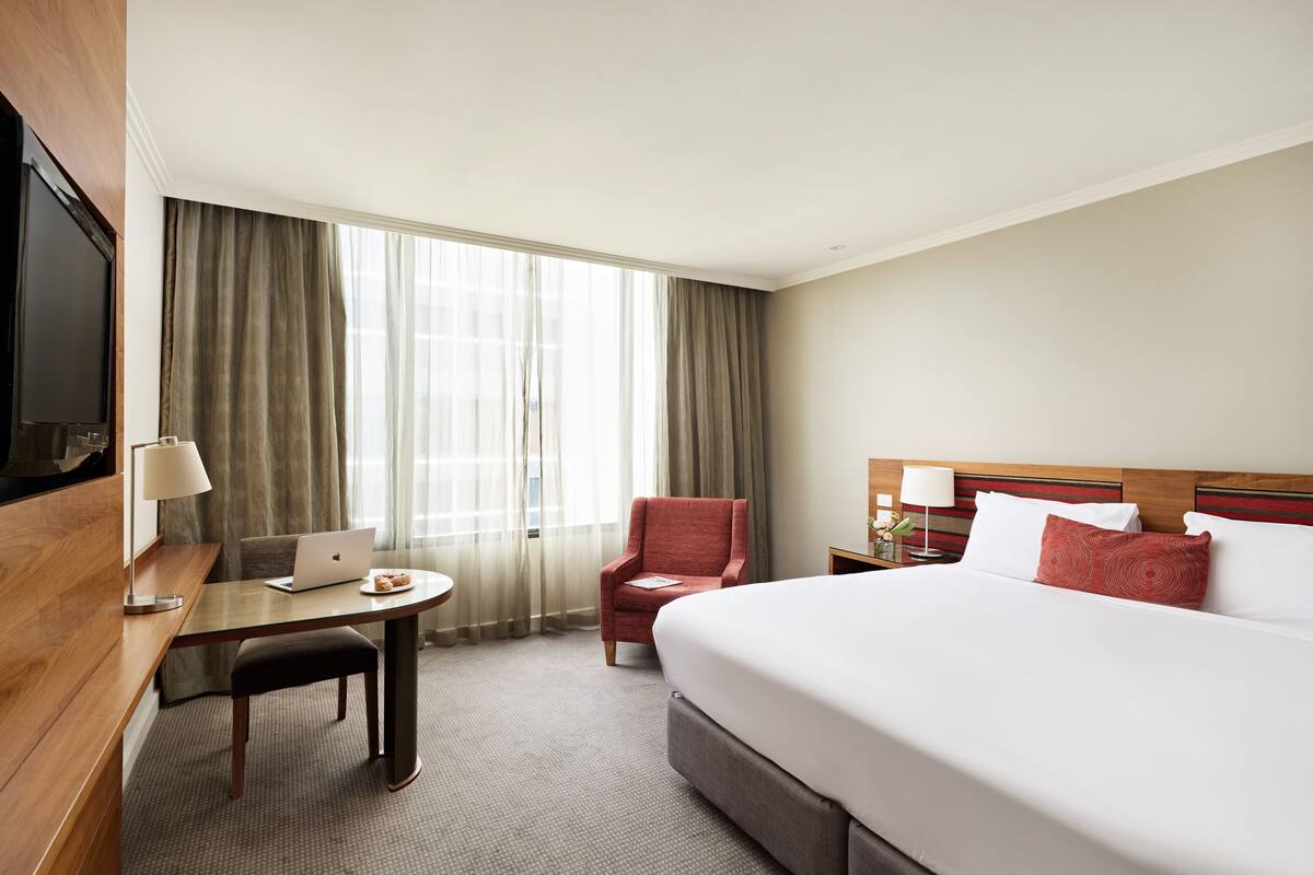 A hotel room with a bed, a desk with a laptop, a chair, a TV, and a window with curtains.
