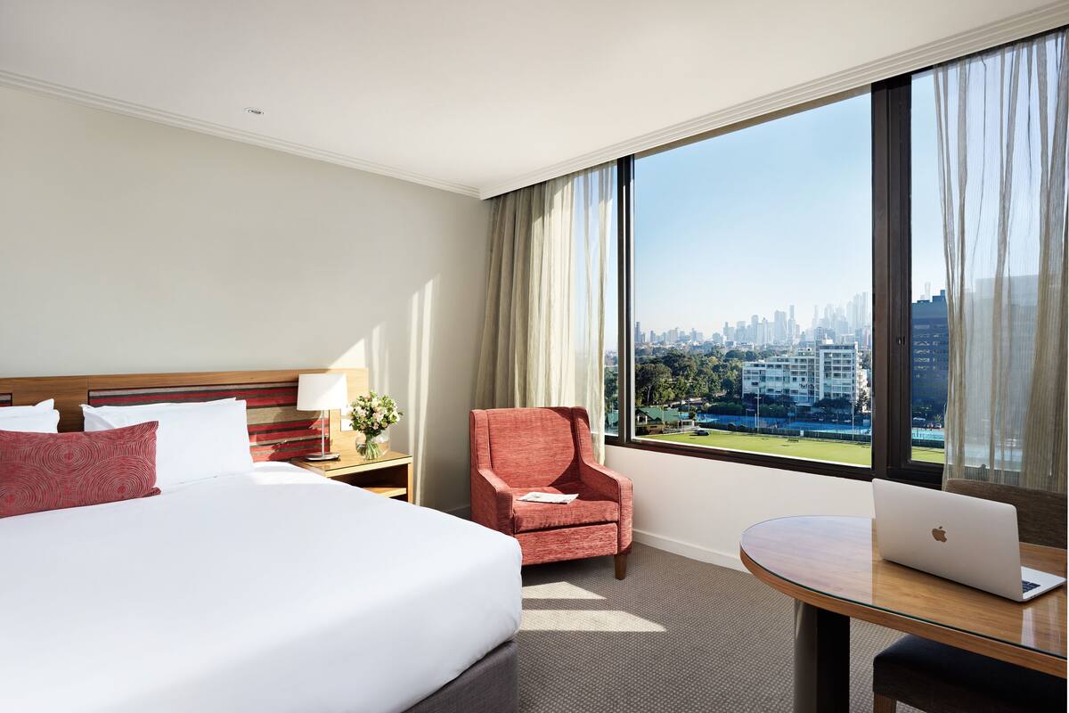 A hotel room with a large bed, a desk with a laptop, a chair, and a view of a city skyline through large windows.