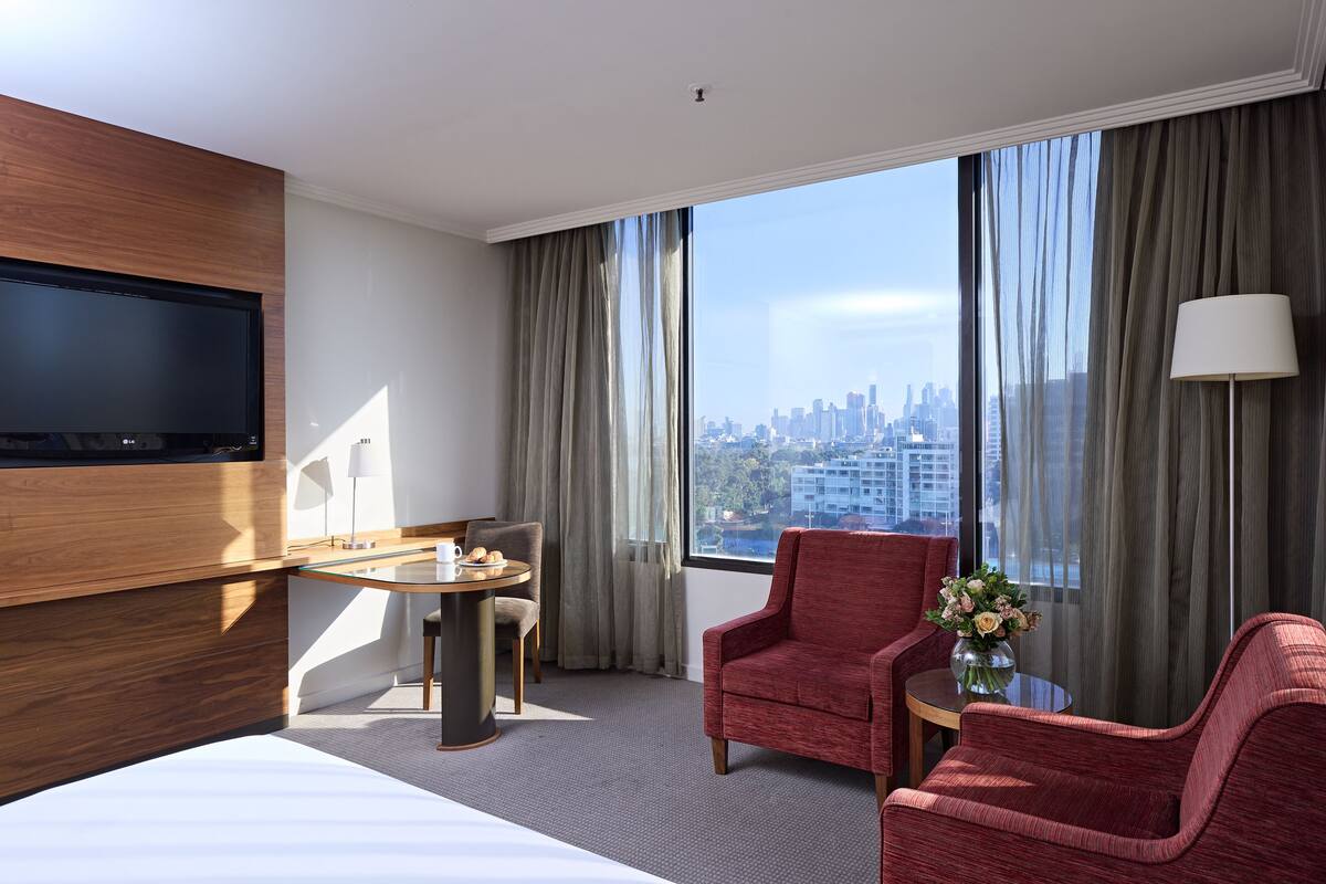 A hotel room with a large bed, two red armchairs, a small round table with a lamp, and a TV mounted on the wall.