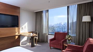 Habitación de hotel con una cama grande, dos sillones rojos, una mesita redonda con una lámpara y un televisor montado en la pared.