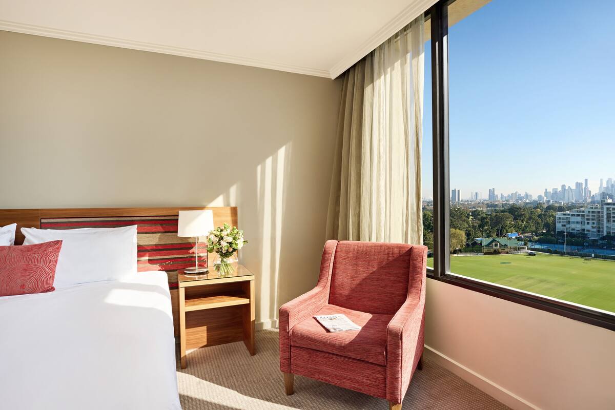 A hotel room with a bed, a nightstand, a chair, and a large window offering a city view.