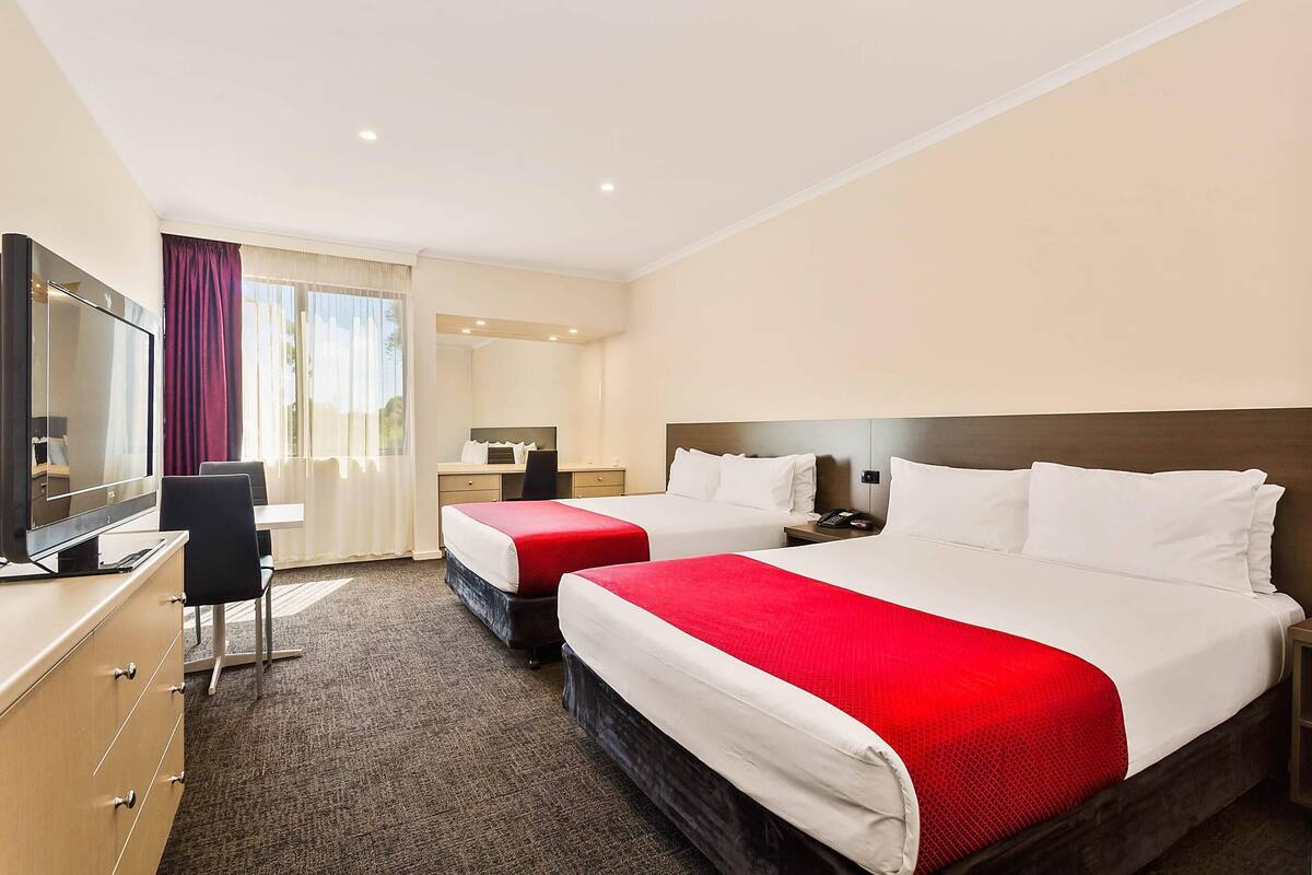 A hotel room with two beds, a desk, a TV, and a window with curtains.