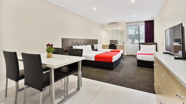 Una habitación de hotel moderna con una cama grande, un sofá cama independiente, un comedor con mesa y sillas, un televisor y una ventana con cortinas.