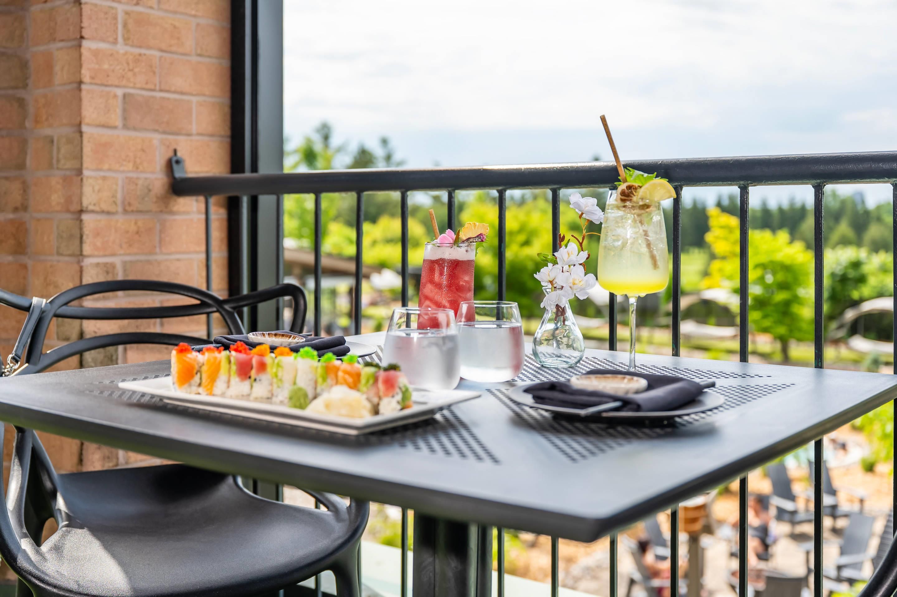 Mesa com sushi, um cocktail e um copo de água numa varanda com vista para vegetação.