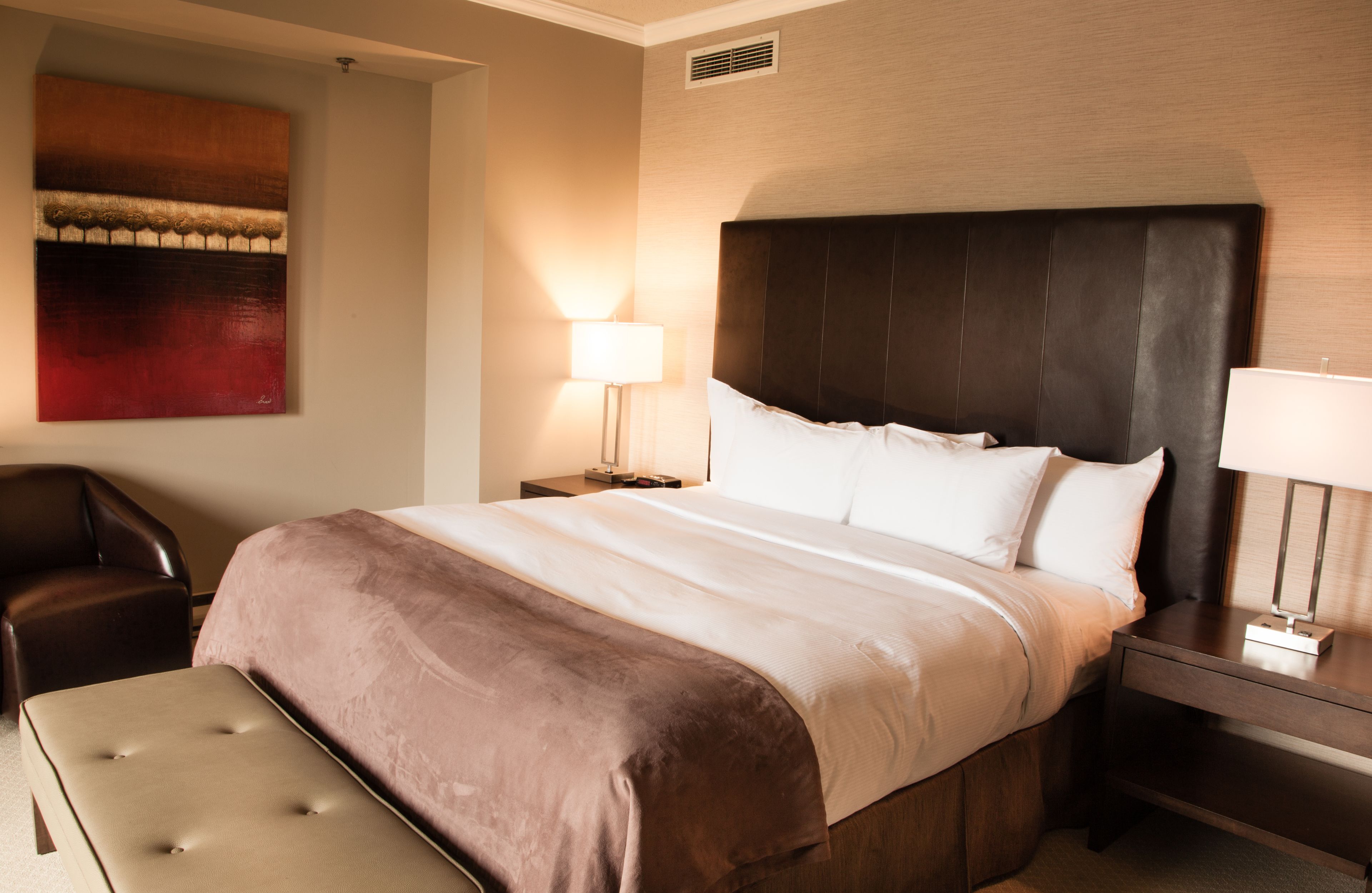 A hotel room with a large bed, a nightstand with a lamp, a chair, and a wall-mounted artwork.