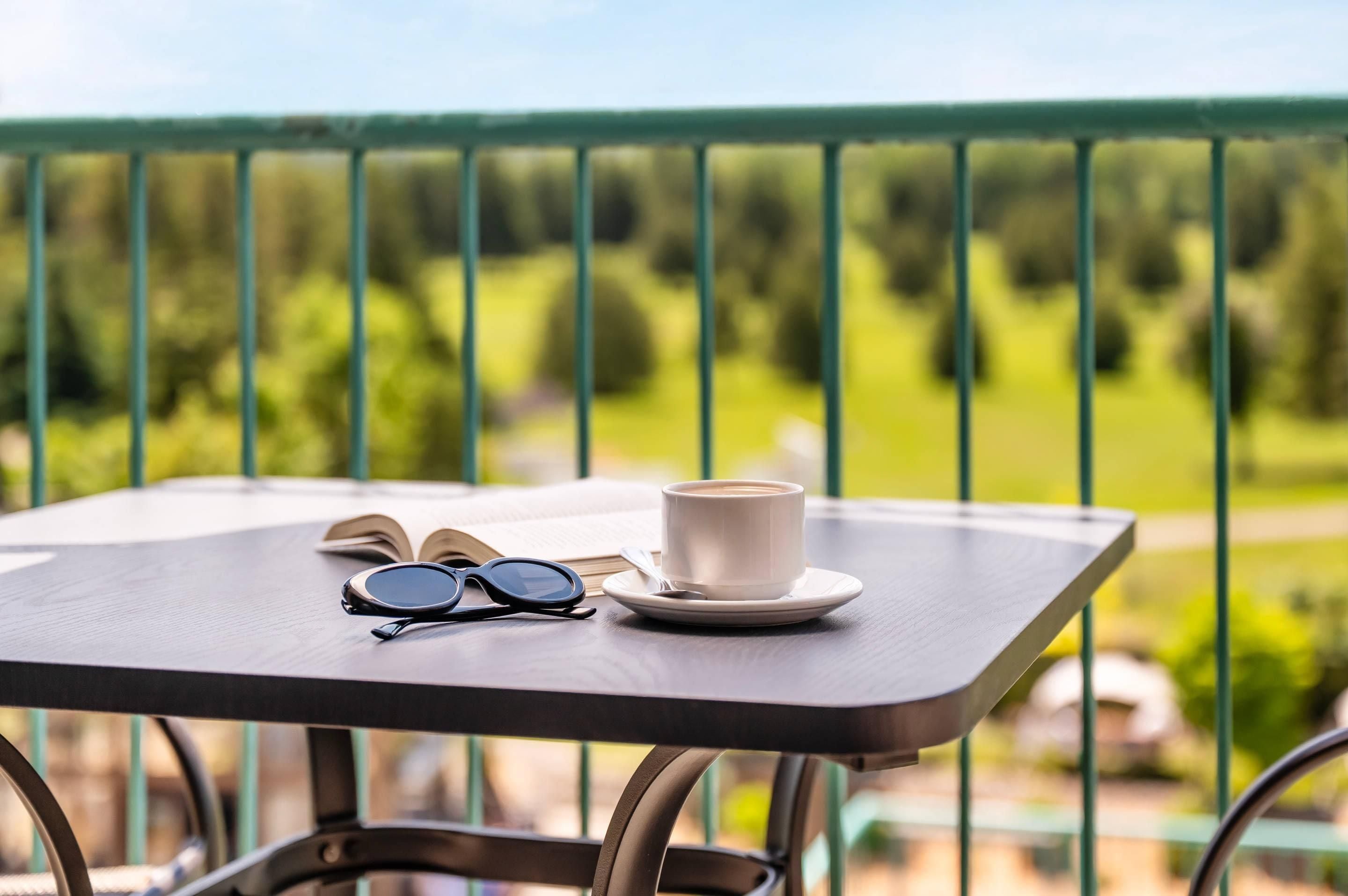 A table with a cup, a pair of glasses, and an open book on a balcony with a green railing and a view of greenery.