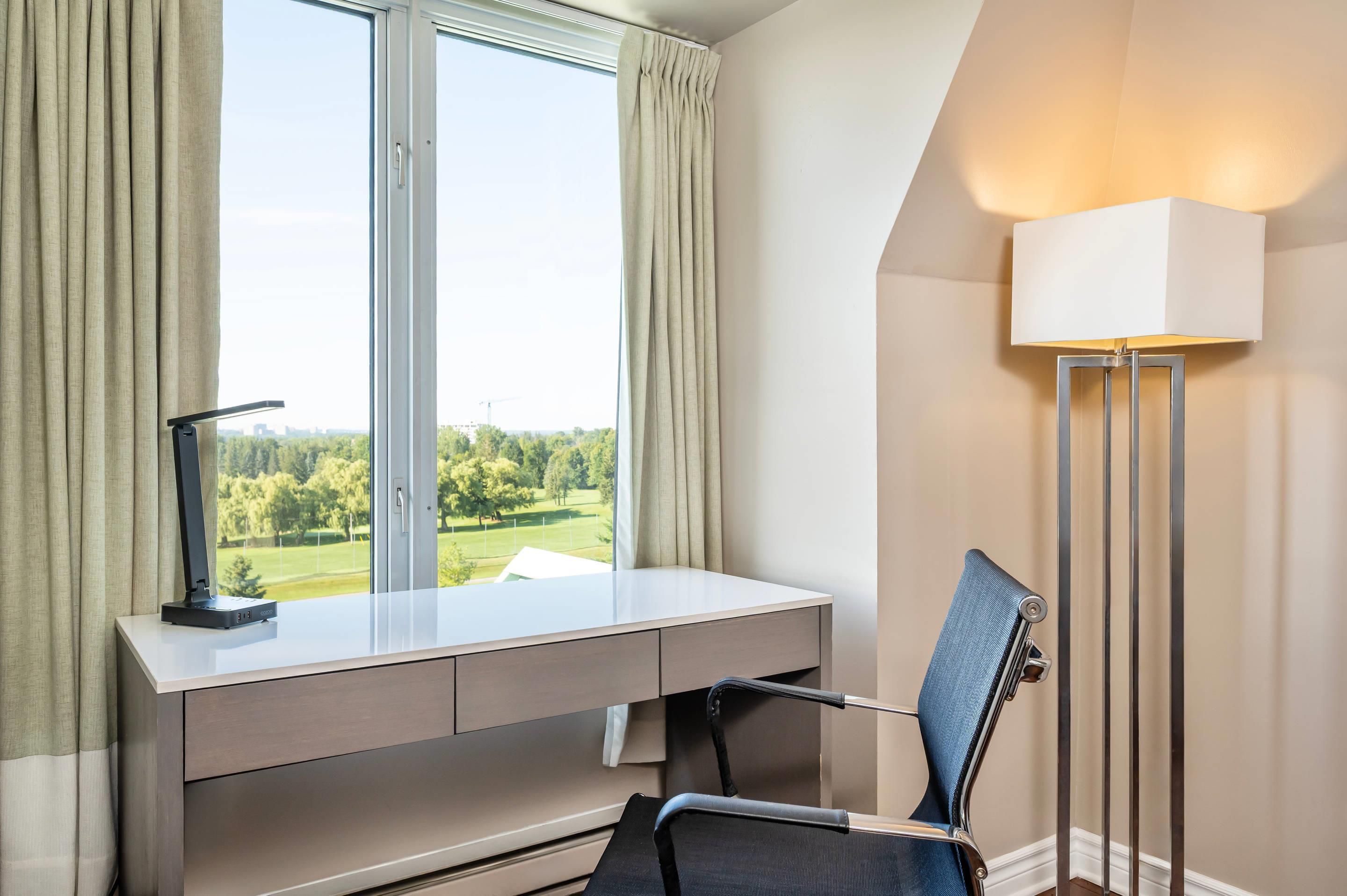 A room with a desk, a chair, a lamp, and a view of a green landscape through a window.