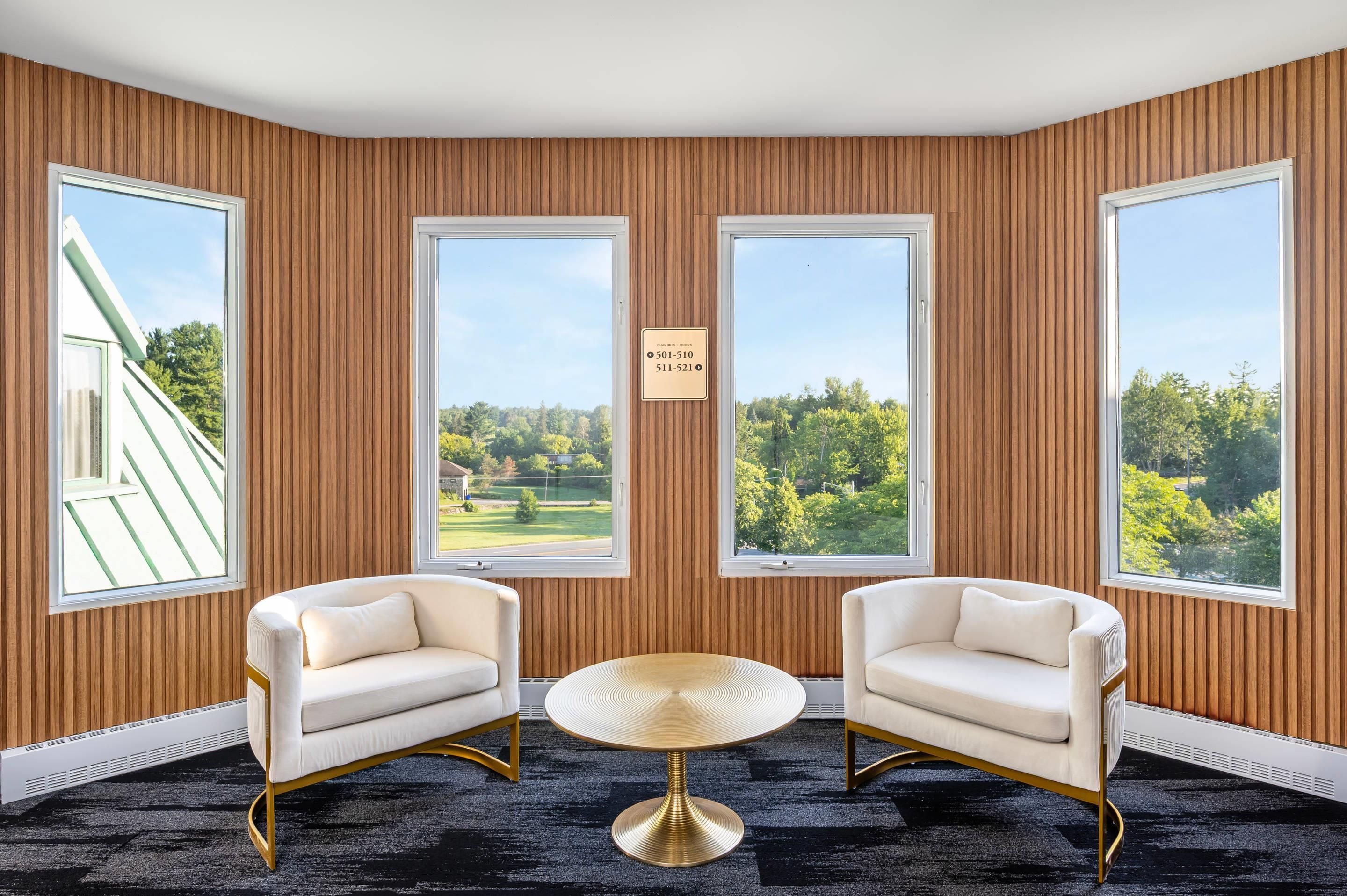 Une pièce avec deux fauteuils blancs, une table ronde dorée et de grandes fenêtres donnant sur des arbres et un terrain de golf.