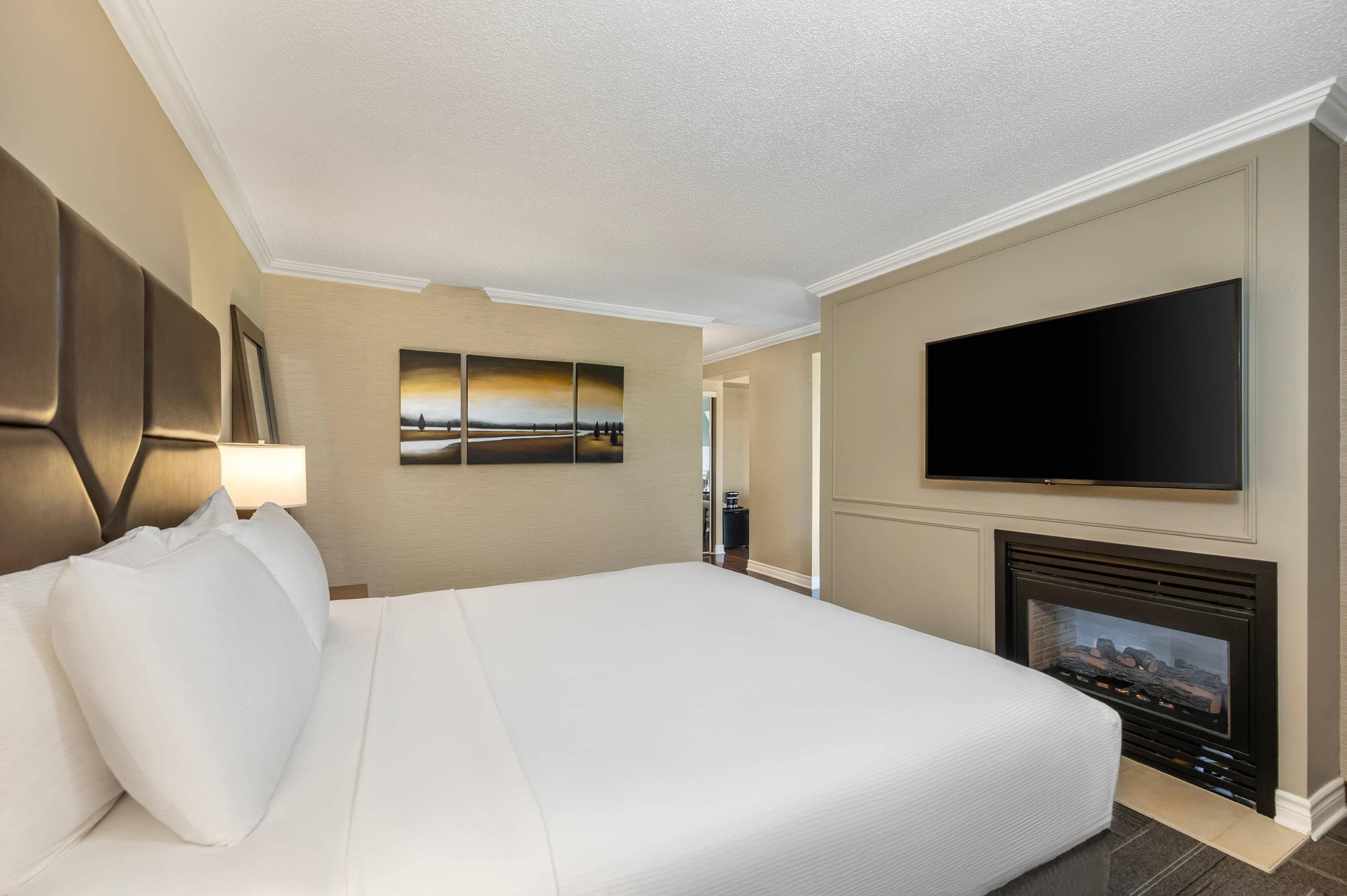 A hotel room with a large bed, a flat-screen TV mounted above a fireplace, and a painting on the wall.