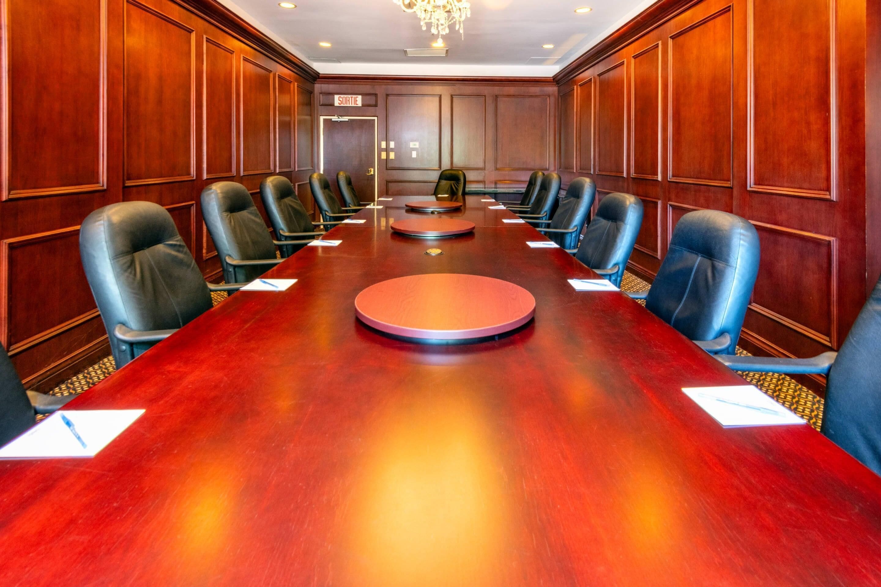 A conference room with a long wooden table, black leather chairs, and a chandelier.