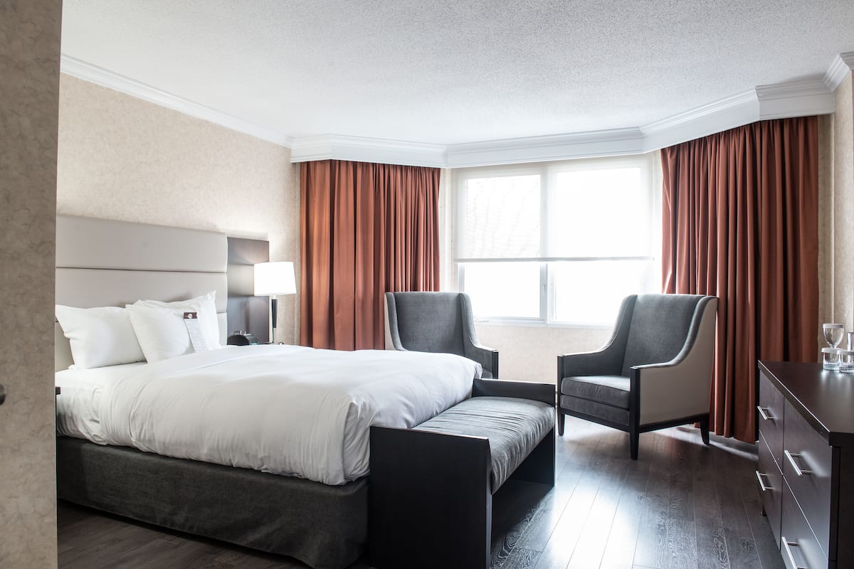 Une chambre d’hôtel avec un lit, deux fauteuils, une commode et une fenêtre avec des rideaux.
