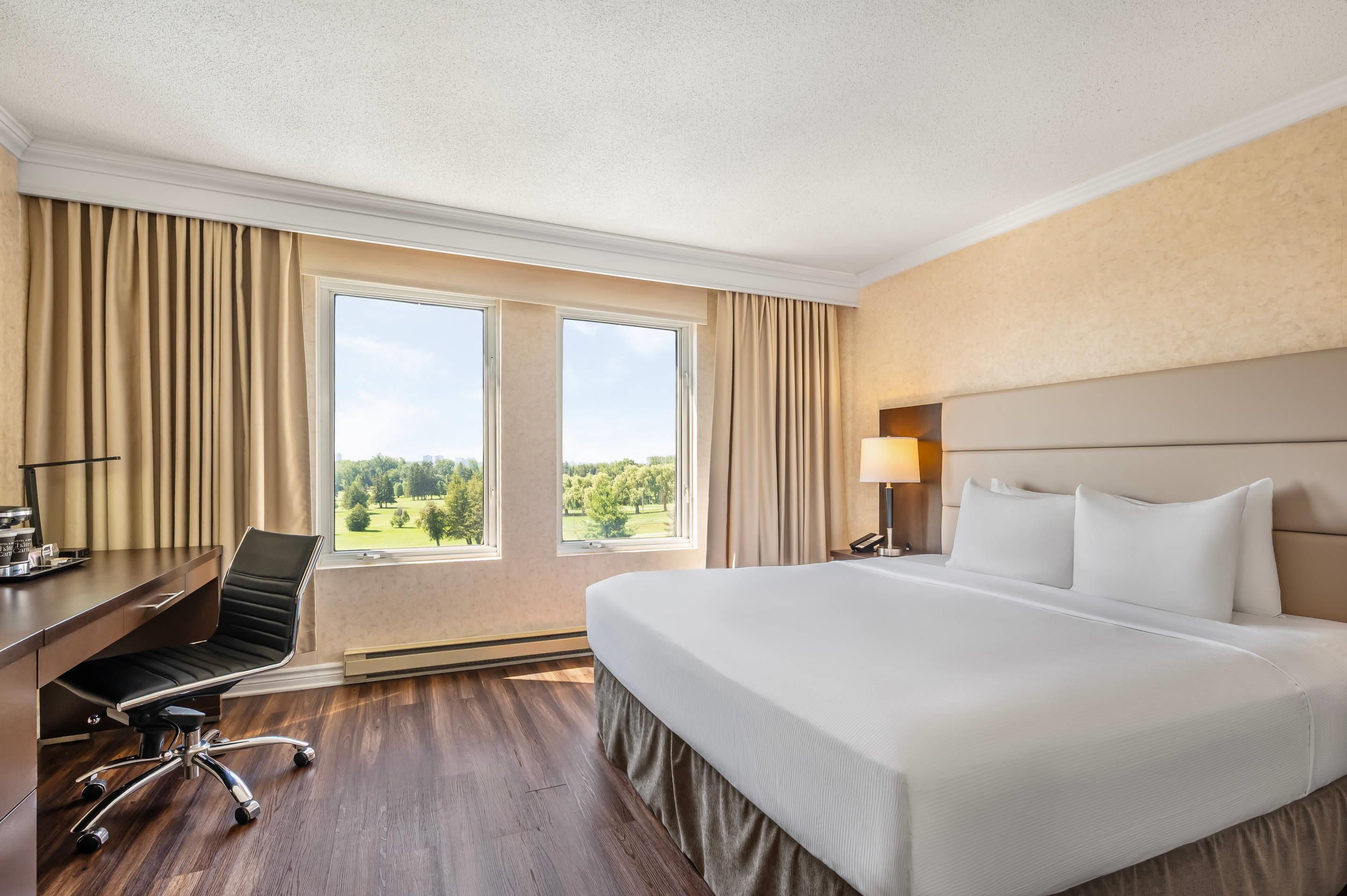 Une chambre d’hôtel avec un grand lit, un bureau avec une chaise, une lampe et une vue sur un parc depuis les fenêtres.