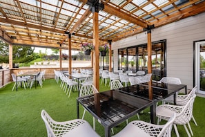 Une terrasse extérieure avec des tables et des chaises, une structure en forme de pergola et une vue sur le jardin environnant.