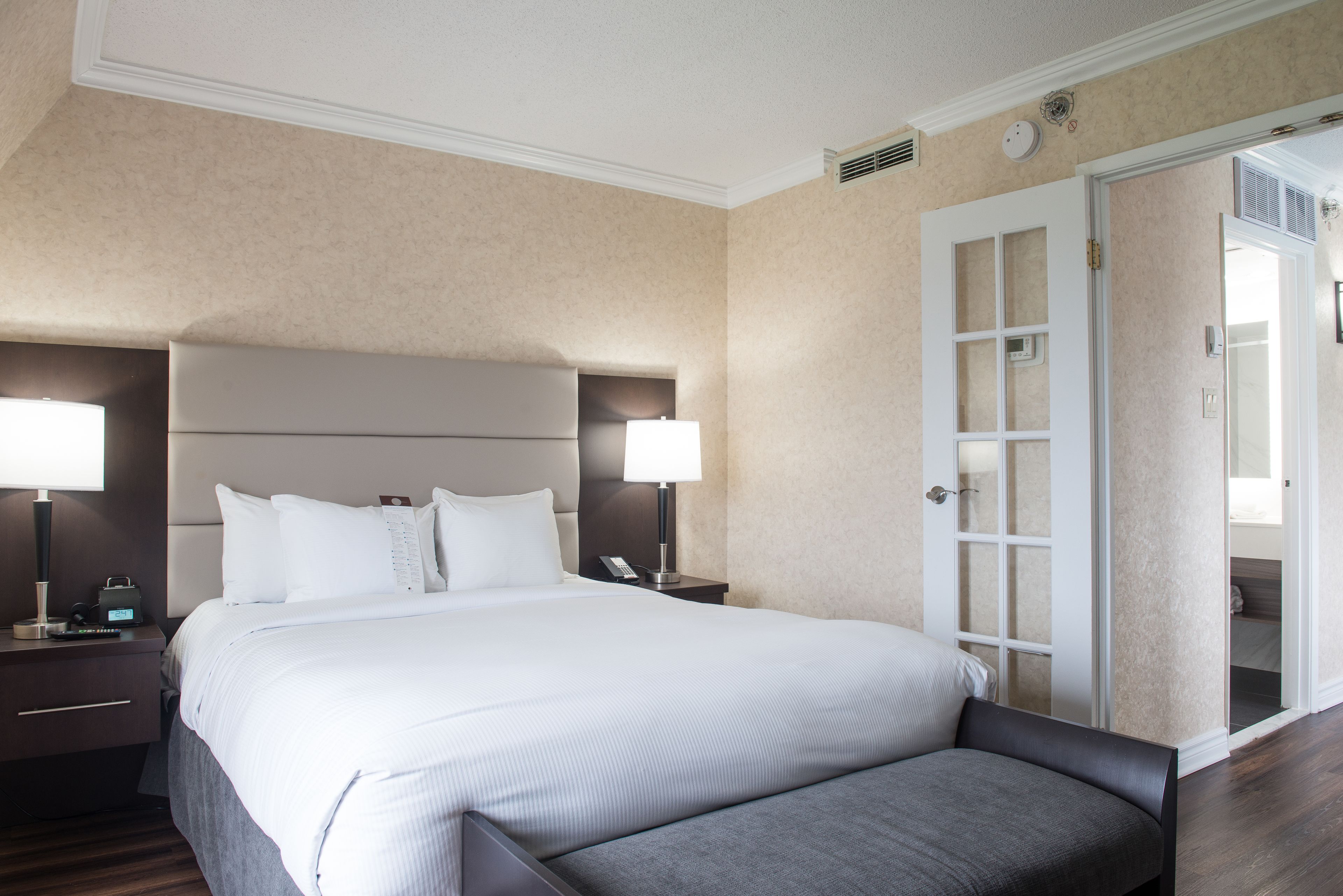 Une chambre d’hôtel avec un grand lit, deux tables de chevet avec des lampes, une tête de lit et une porte donnant sur une salle de bain.