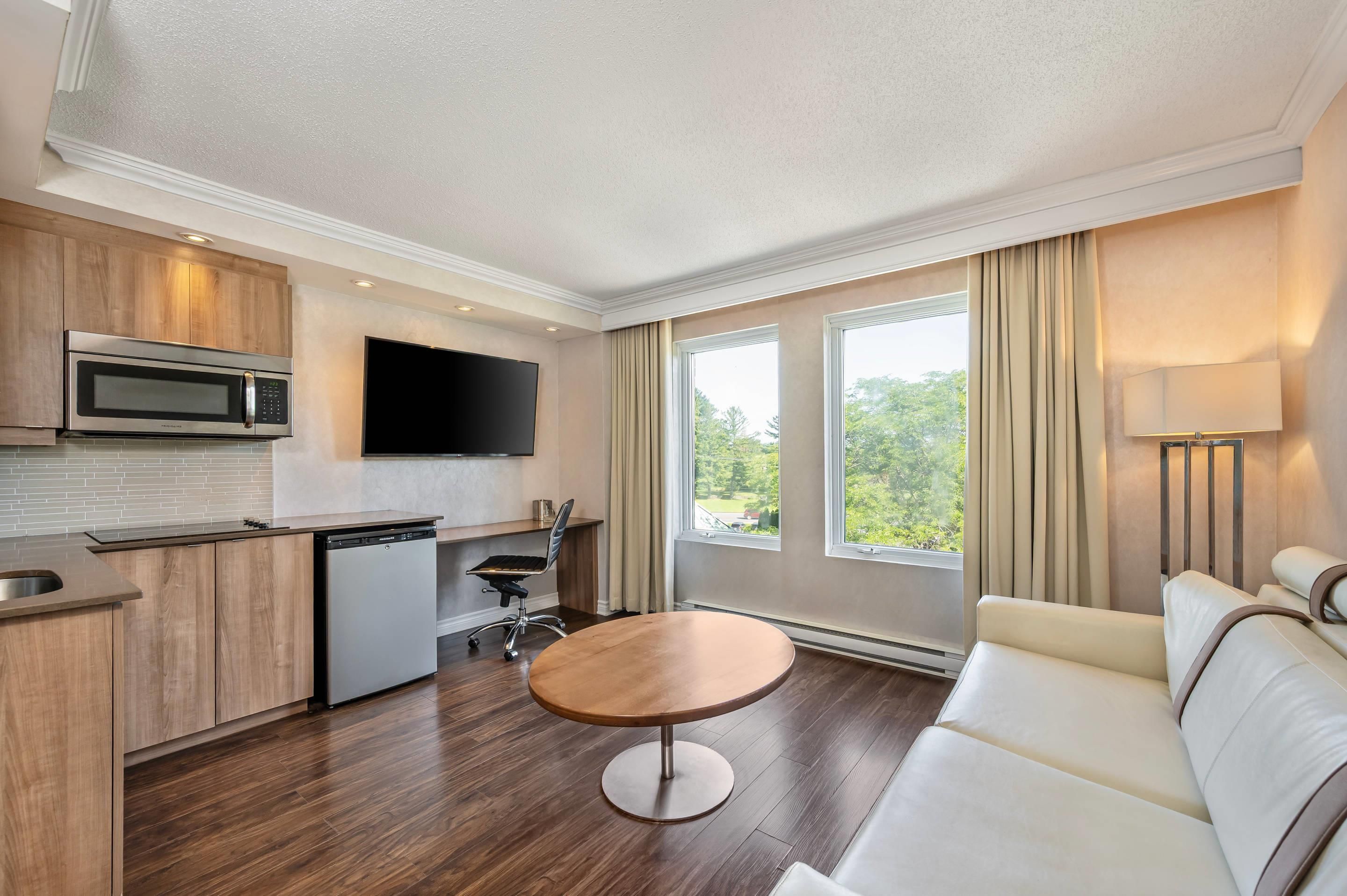 A modern hotel room with a kitchenette, a flat-screen TV, a dining table, and a sofa.