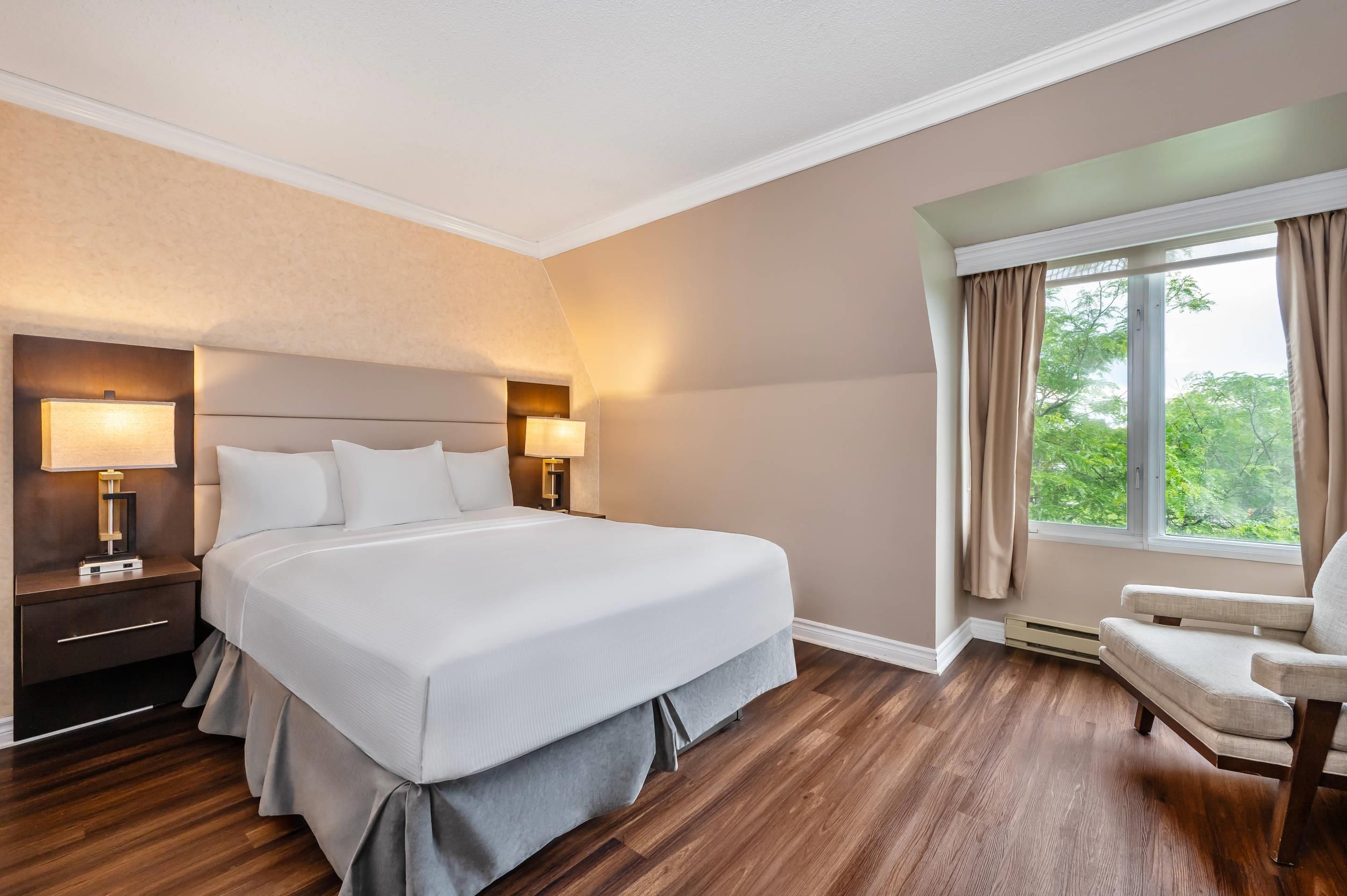 A hotel room with a large bed, bedside lamps, a nightstand, a chair, and a window with curtains.