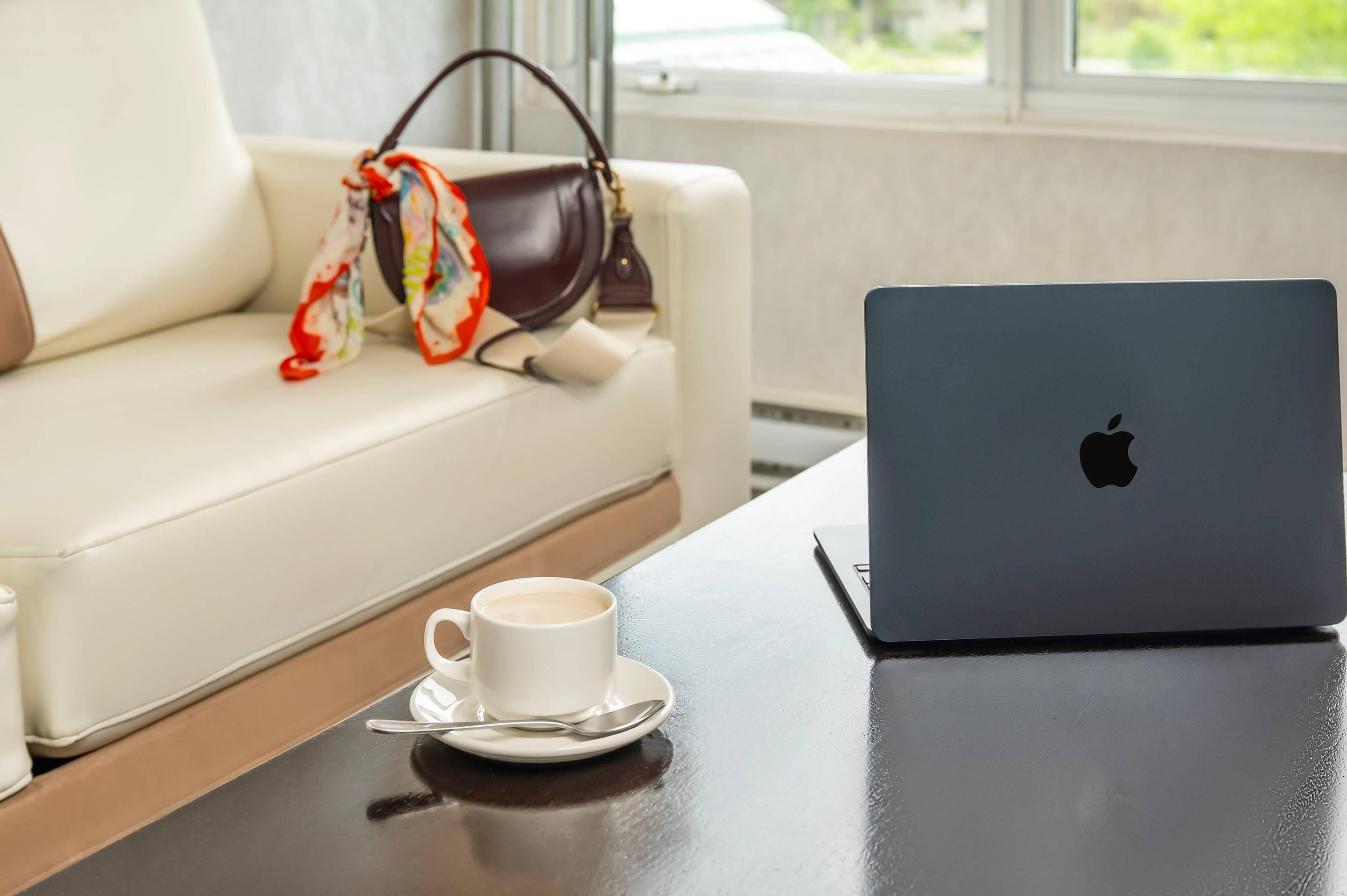 Un portátil sobre una mesa con una taza y platillo, un sofá blanco con un bolso marrón, y una ventana con vistas.