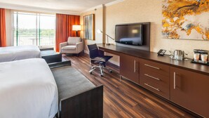 Habitación de hotel con dos camas, un escritorio, un televisor, una silla y un ventanal con cortinas.