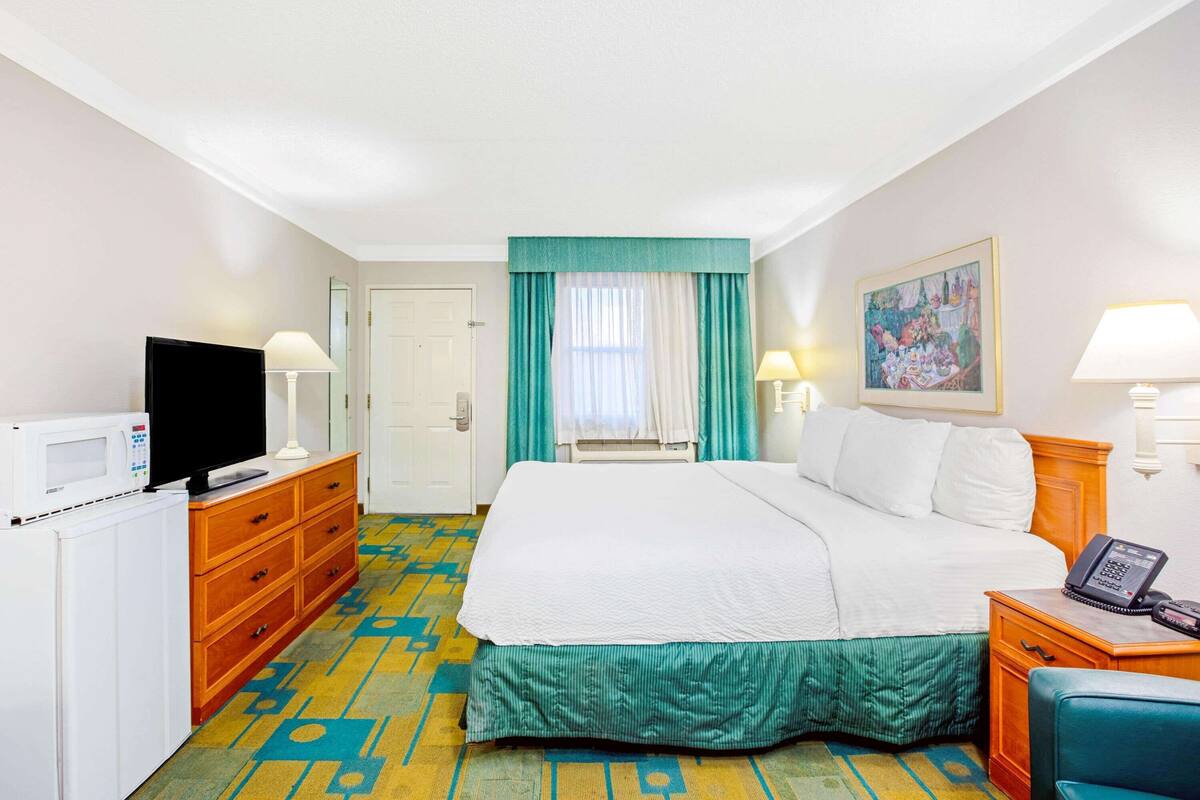 A hotel room with a bed, a TV, a desk with a lamp, a telephone, and a painting on the wall.