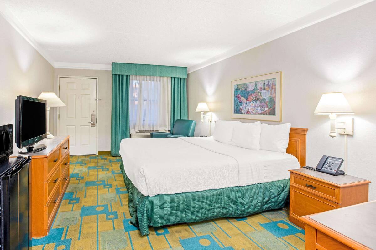 A hotel room with a bed, a desk, a chair, a TV, and a painting on the wall.