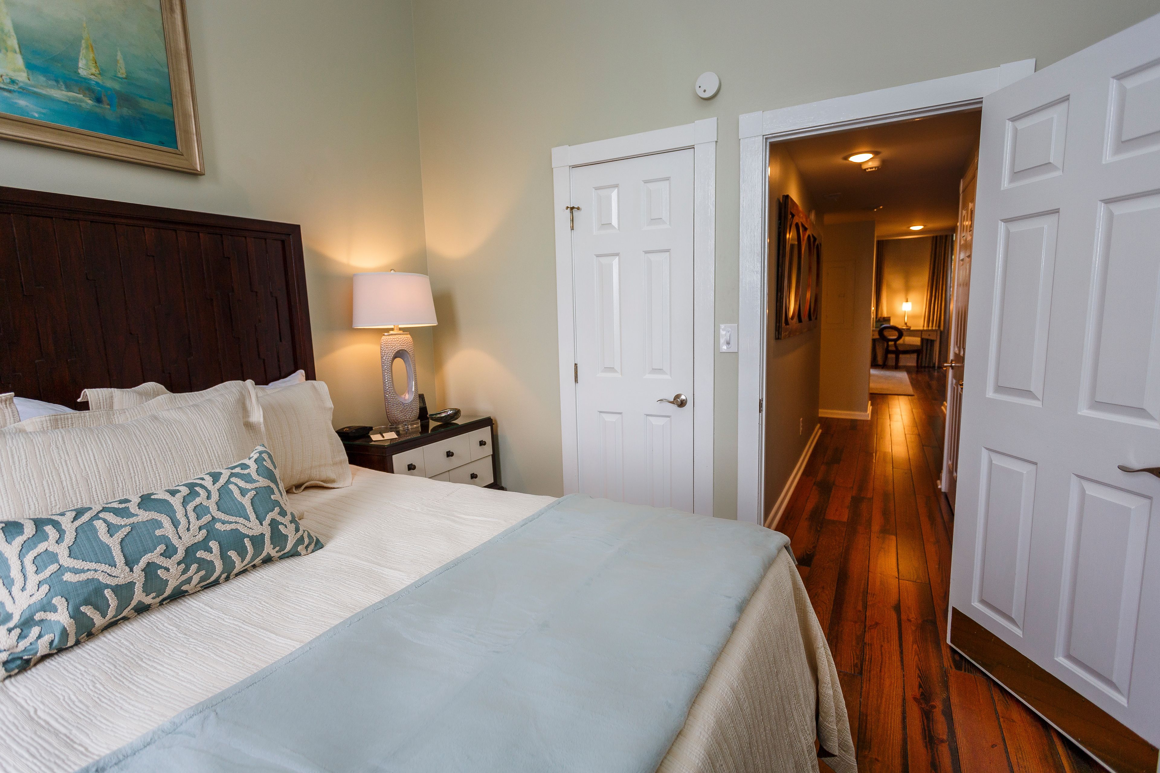 A hotel room with a bed, nightstand, lamp, and a hallway leading to another room.