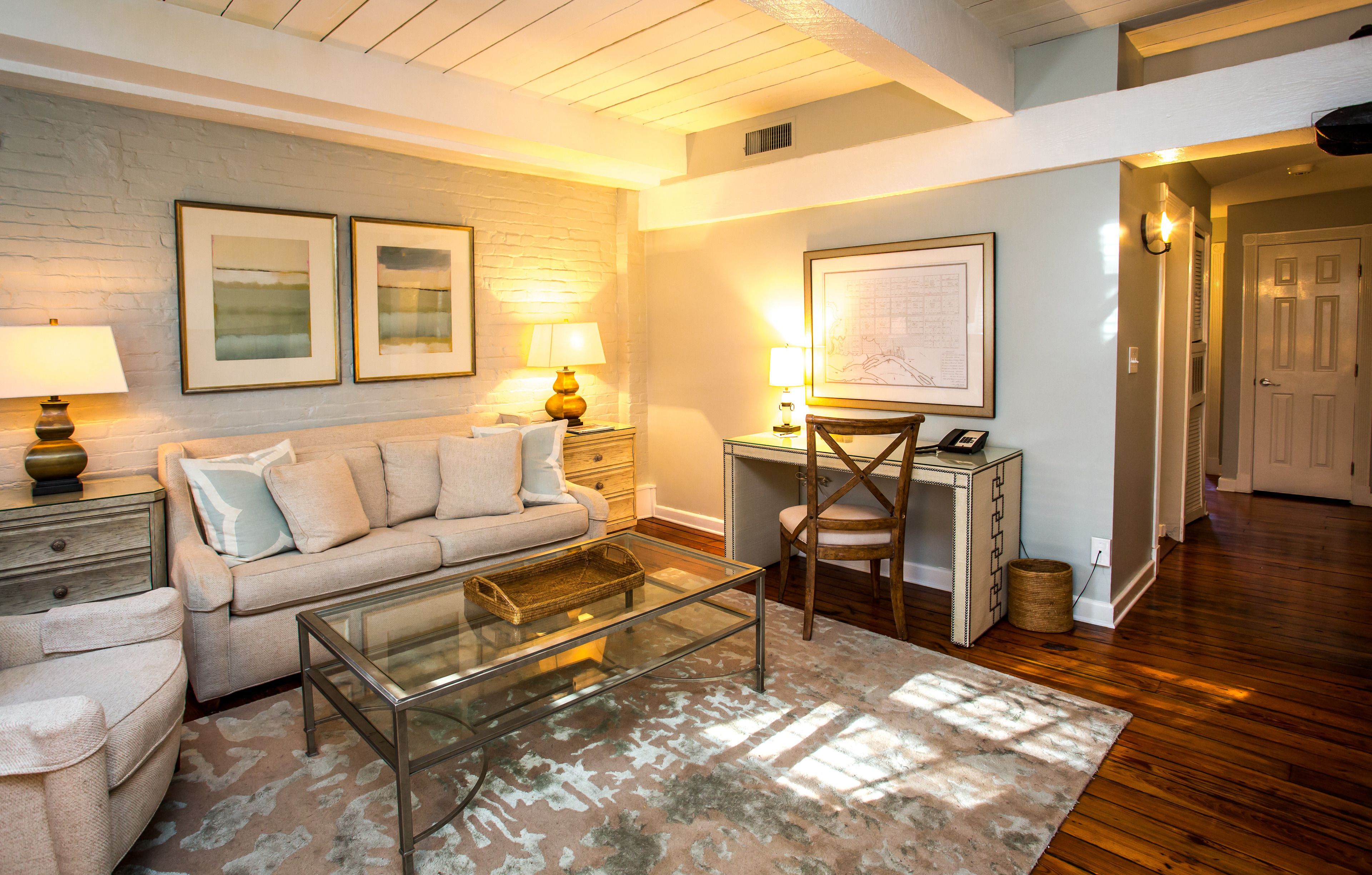A living room with a sofa, coffee table, and lamps. There is a desk with a chair and a framed map on the wall.