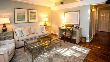 A living room with a sofa, coffee table, and lamps. There is a desk with a chair and a framed map on the wall.