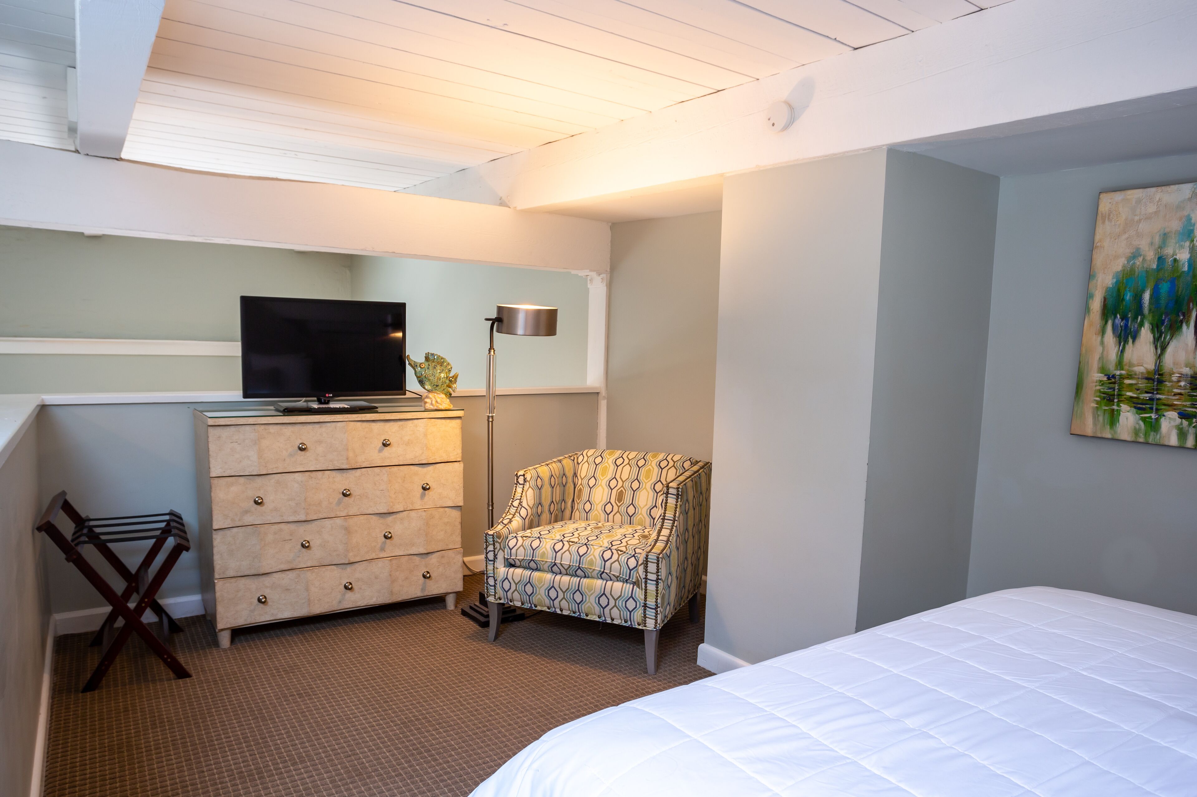 Un dormitorio con cama, cómoda, silla, televisor y un cuadro en la pared.