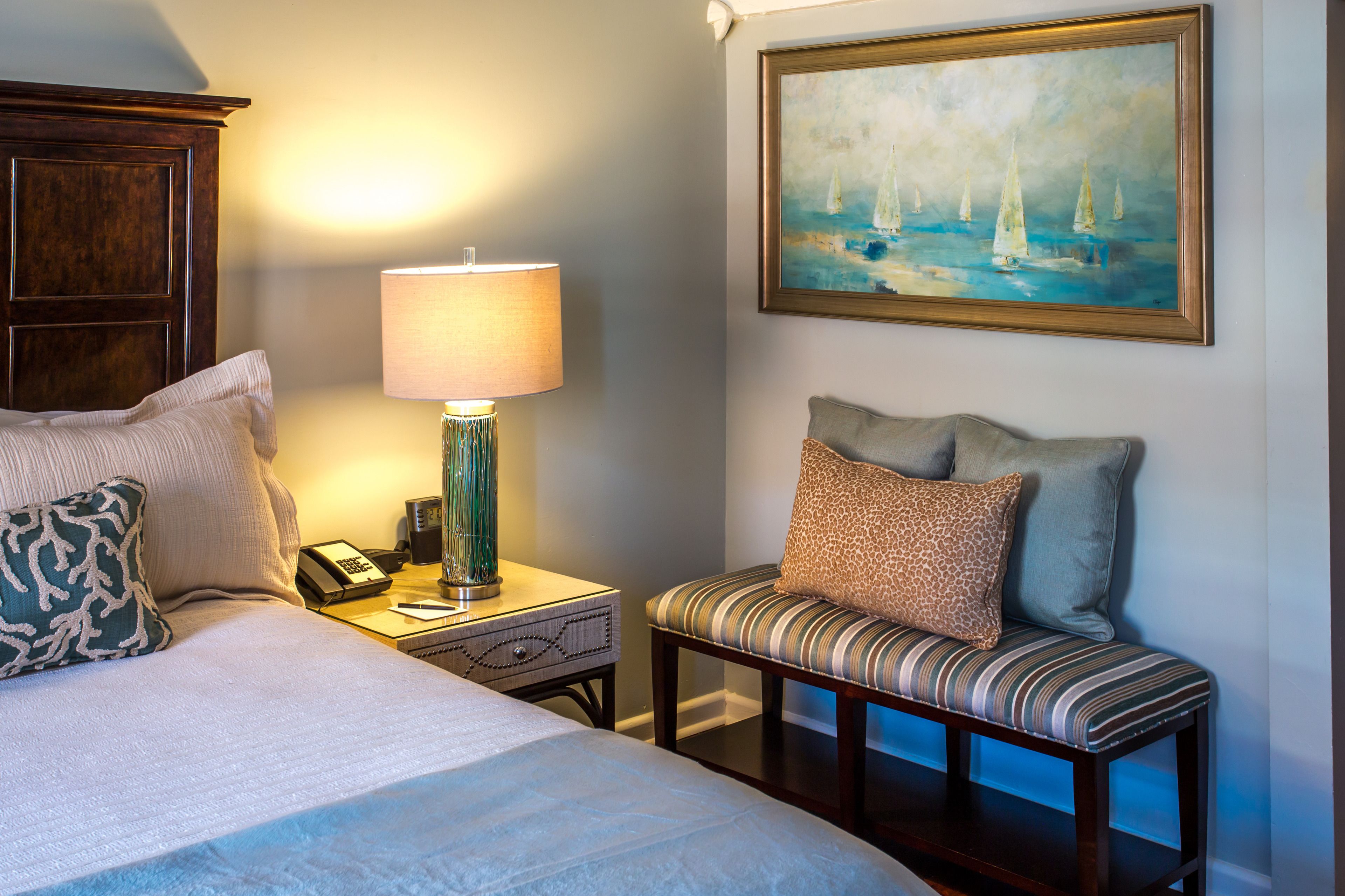 A hotel room with a bed, bedside lamp, a bench with cushions, a painting of sailboats, and a telephone on the nightstand.