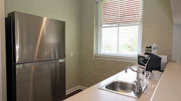 Una cocina con refrigerador de acero inoxidable, una ventana con persianas, un lavamanos con grifo antiguo y un microondas.