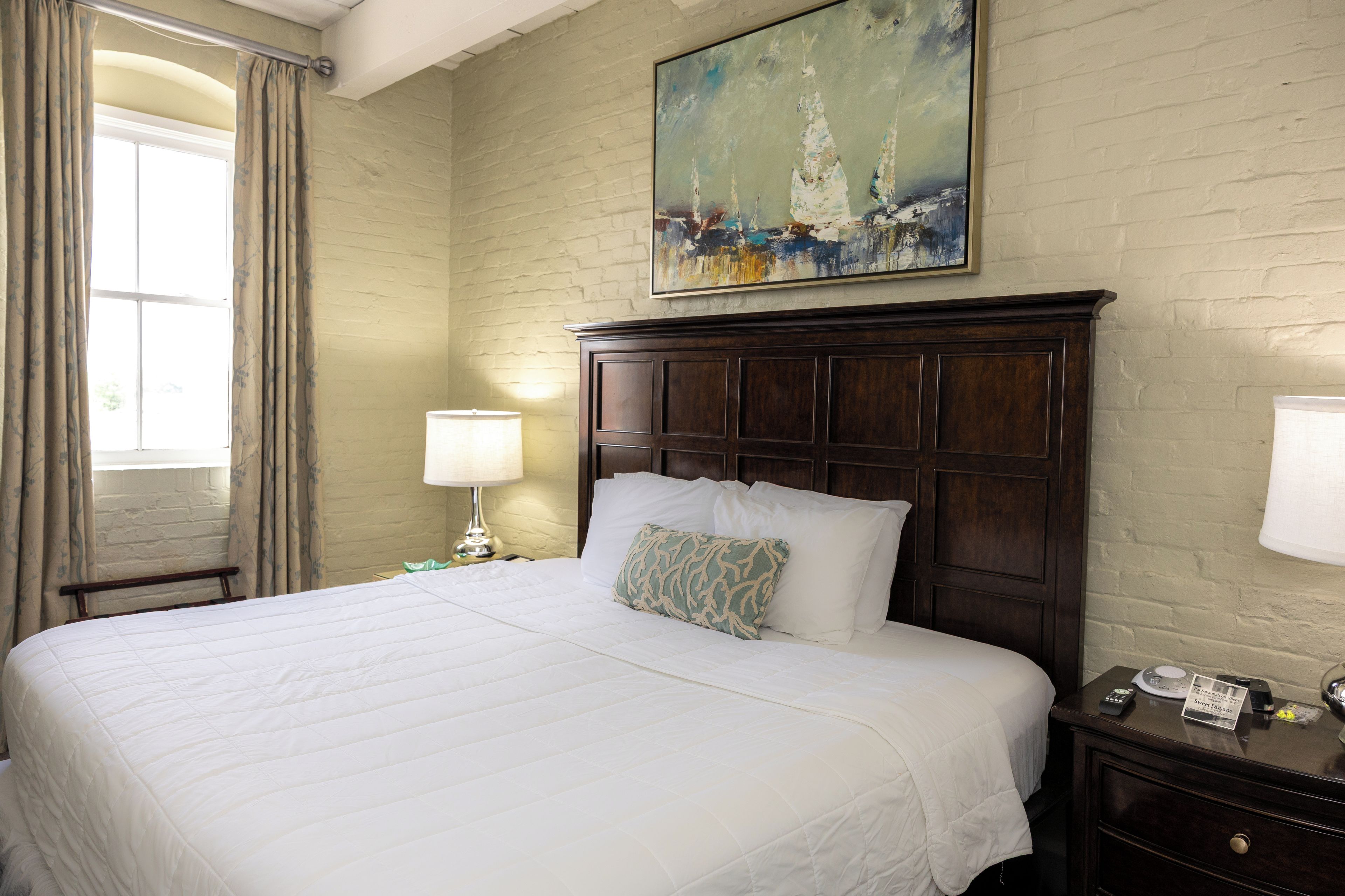 Une chambre d’hôtel avec un grand lit, une table de chevet avec une lampe, un tableau au-dessus du lit et une fenêtre avec des rideaux.