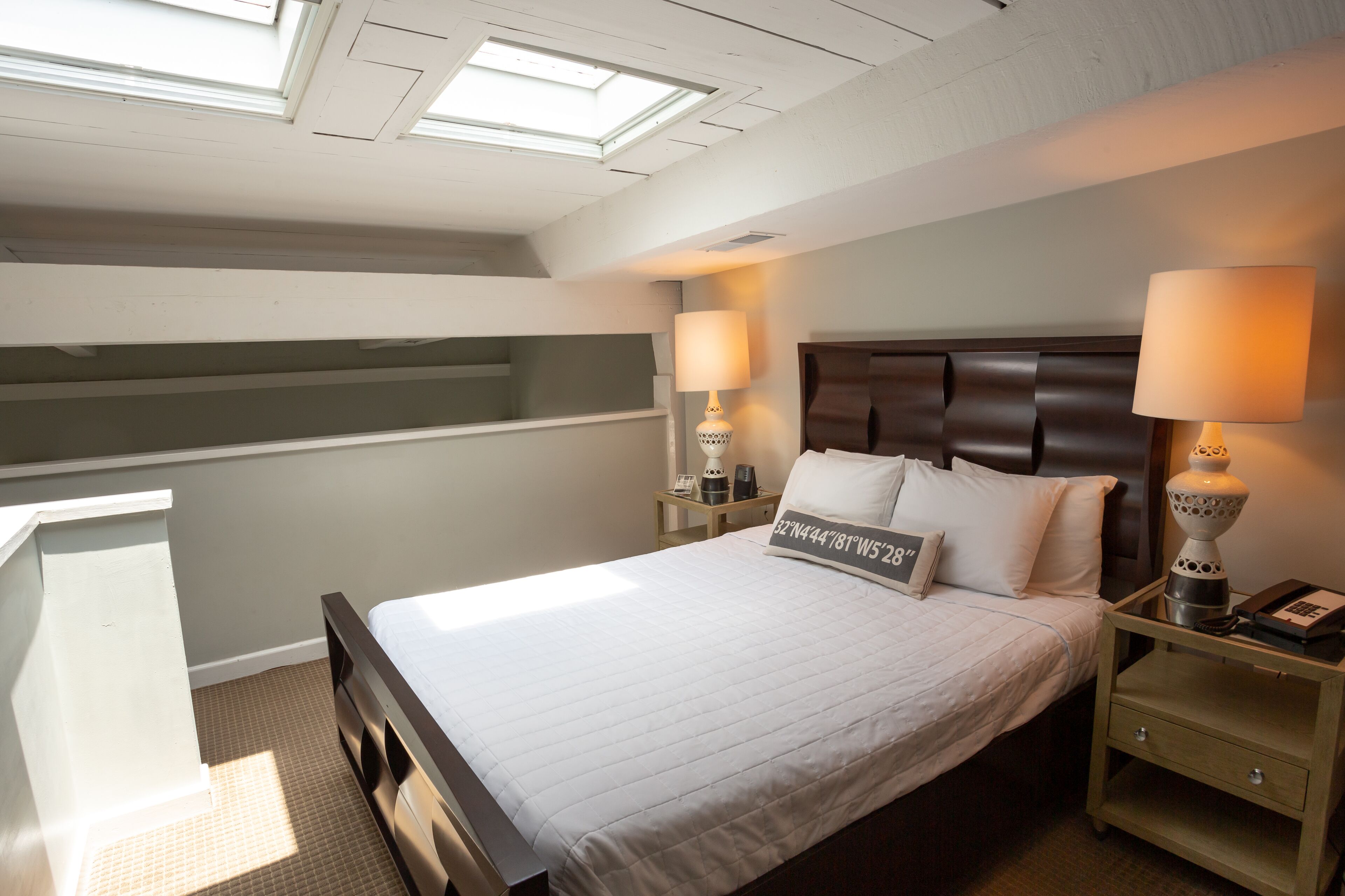 A bedroom with a bed, two bedside tables with lamps, a headboard, a small desk, and a skylight.