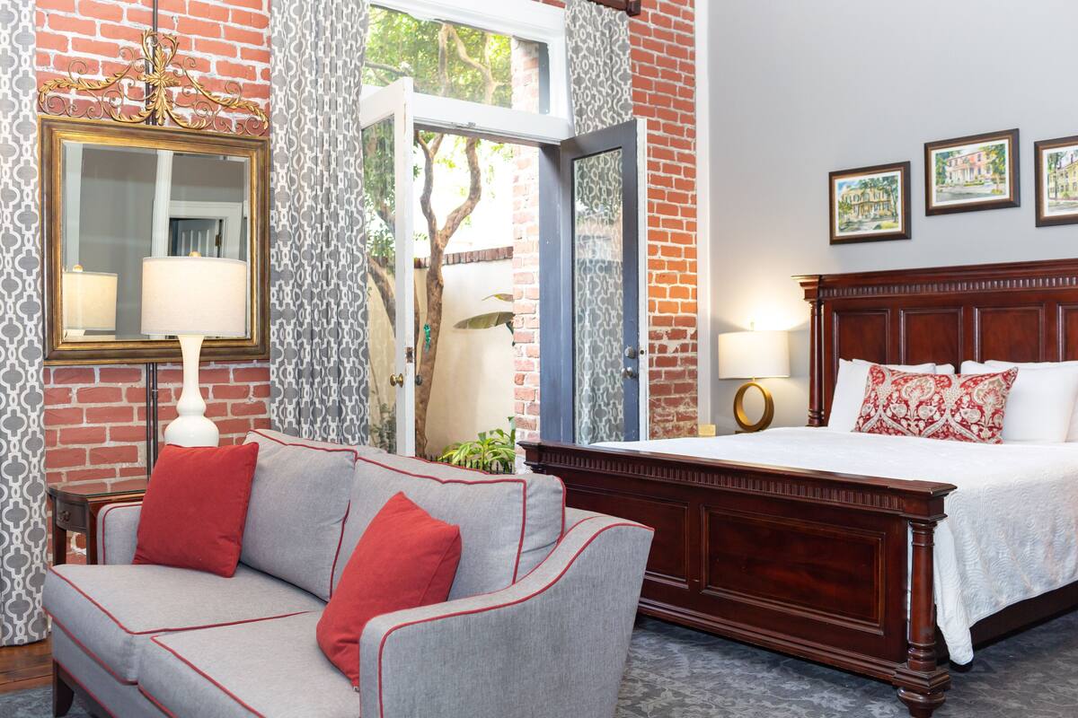 A hotel room with a bed, a sofa, a mirror, and a window with a view of trees.