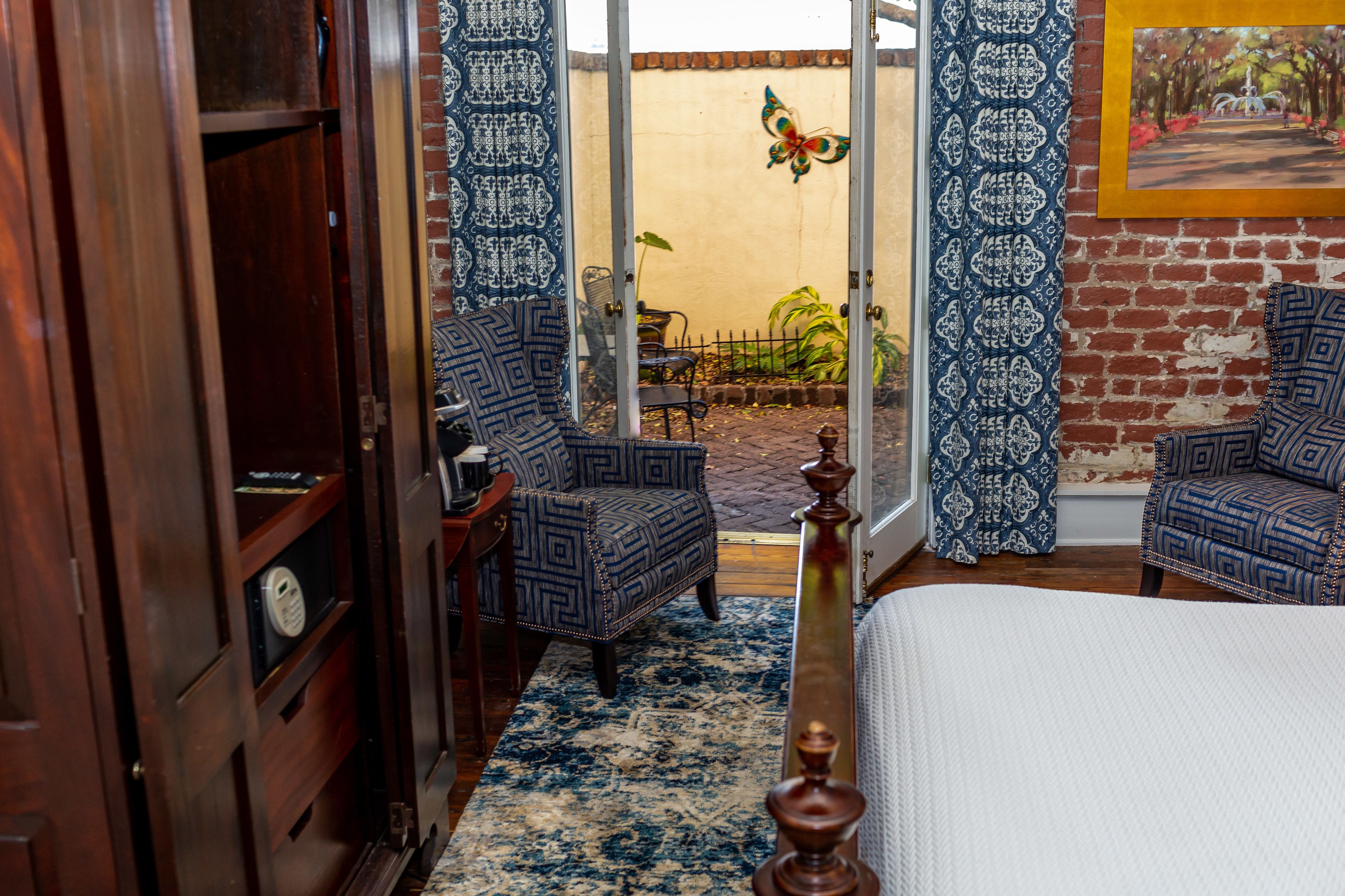 Una habitación con una cama, dos sillones estampados, un armario de madera, un tapiz con diseño y vistas a un jardín exterior a través de un ventanal.