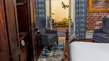 Una habitación con una cama, dos sillones estampados, un armario de madera, un tapiz con diseño y vistas a un jardín exterior a través de un ventanal.