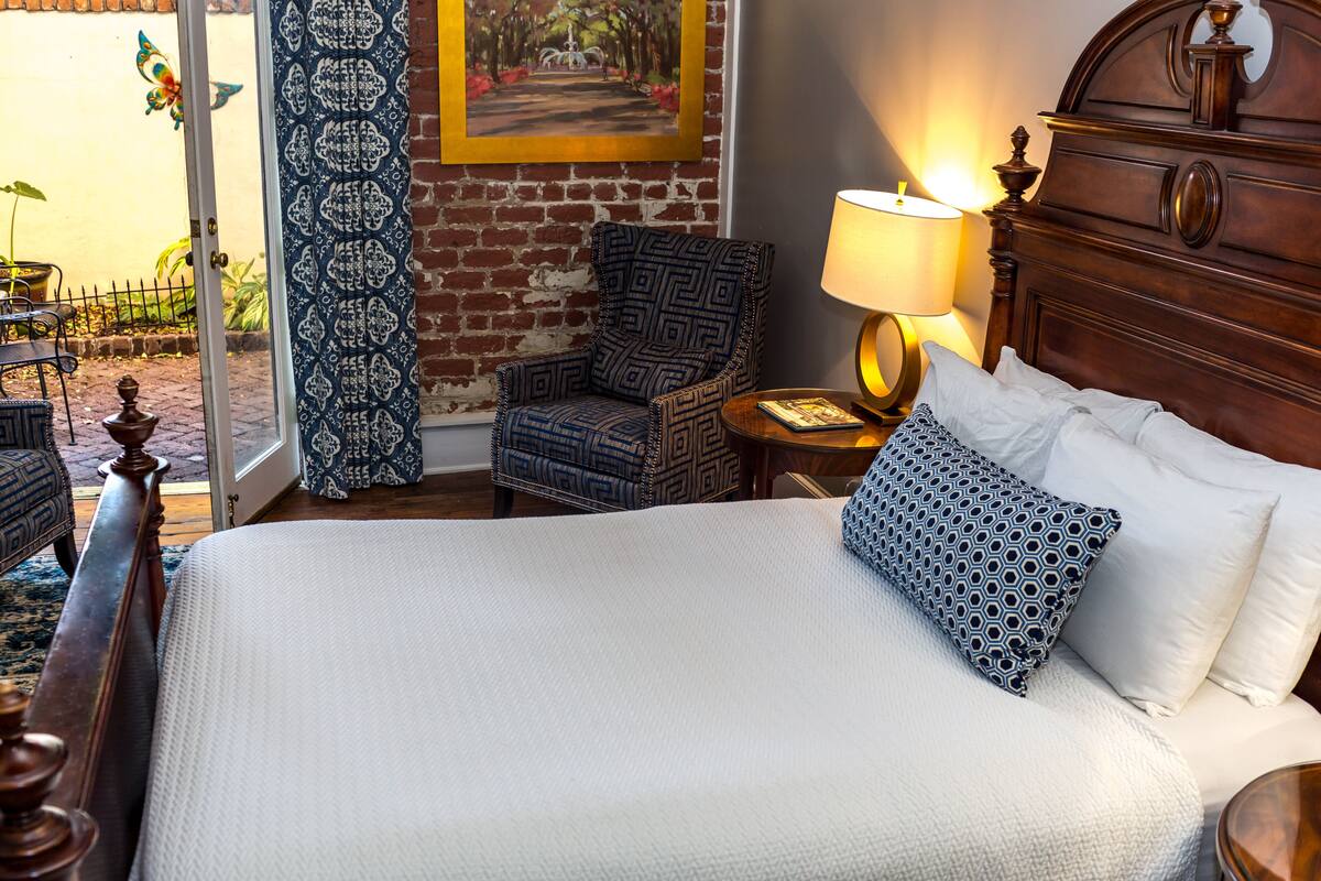 A hotel room with a large bed, a wooden headboard, a patterned armchair, a small table with a lamp, and a view through a glass door.
