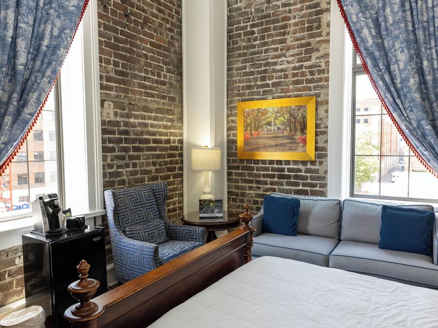 A hotel room with a brick wall, a bed, a sofa, a chair, a lamp, and a painting.