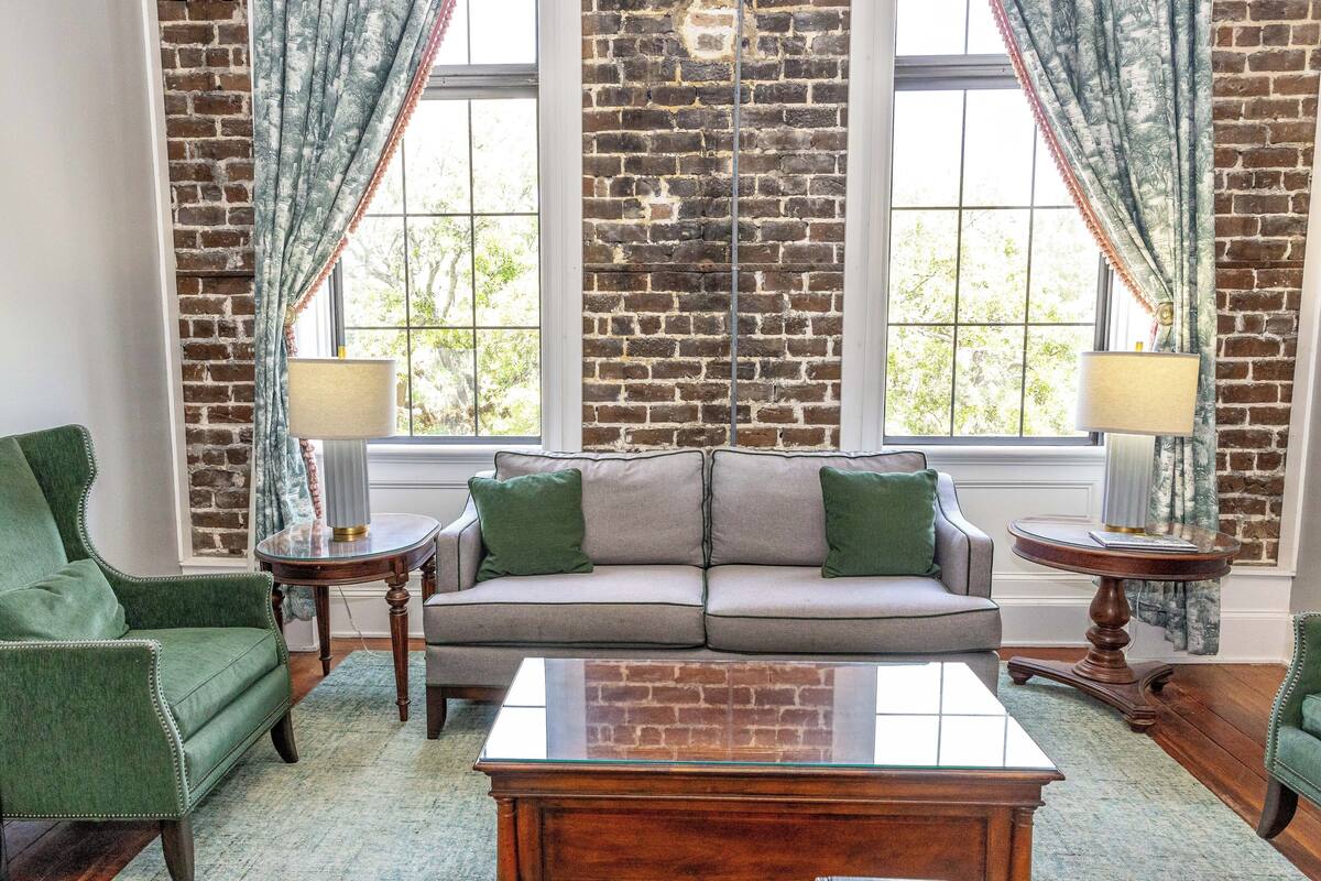 A living room with a brick wall, a gray sofa, green armchairs, and a glass coffee table.