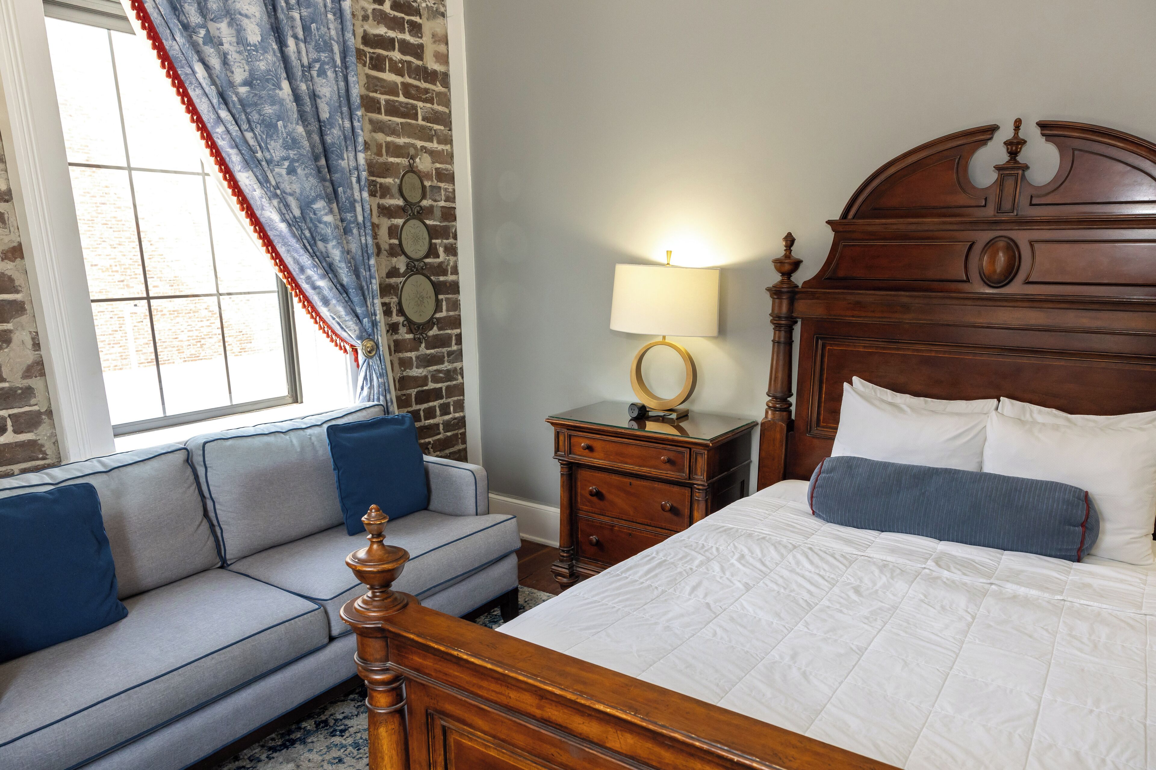 A bedroom with a wooden bed, a grey sofa, a nightstand with a lamp, and a window with curtains.