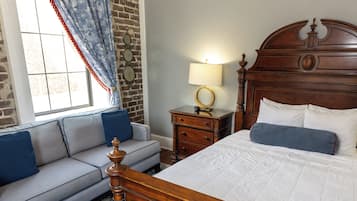 A bedroom with a wooden bed, a grey sofa, a nightstand with a lamp, and a window with curtains.