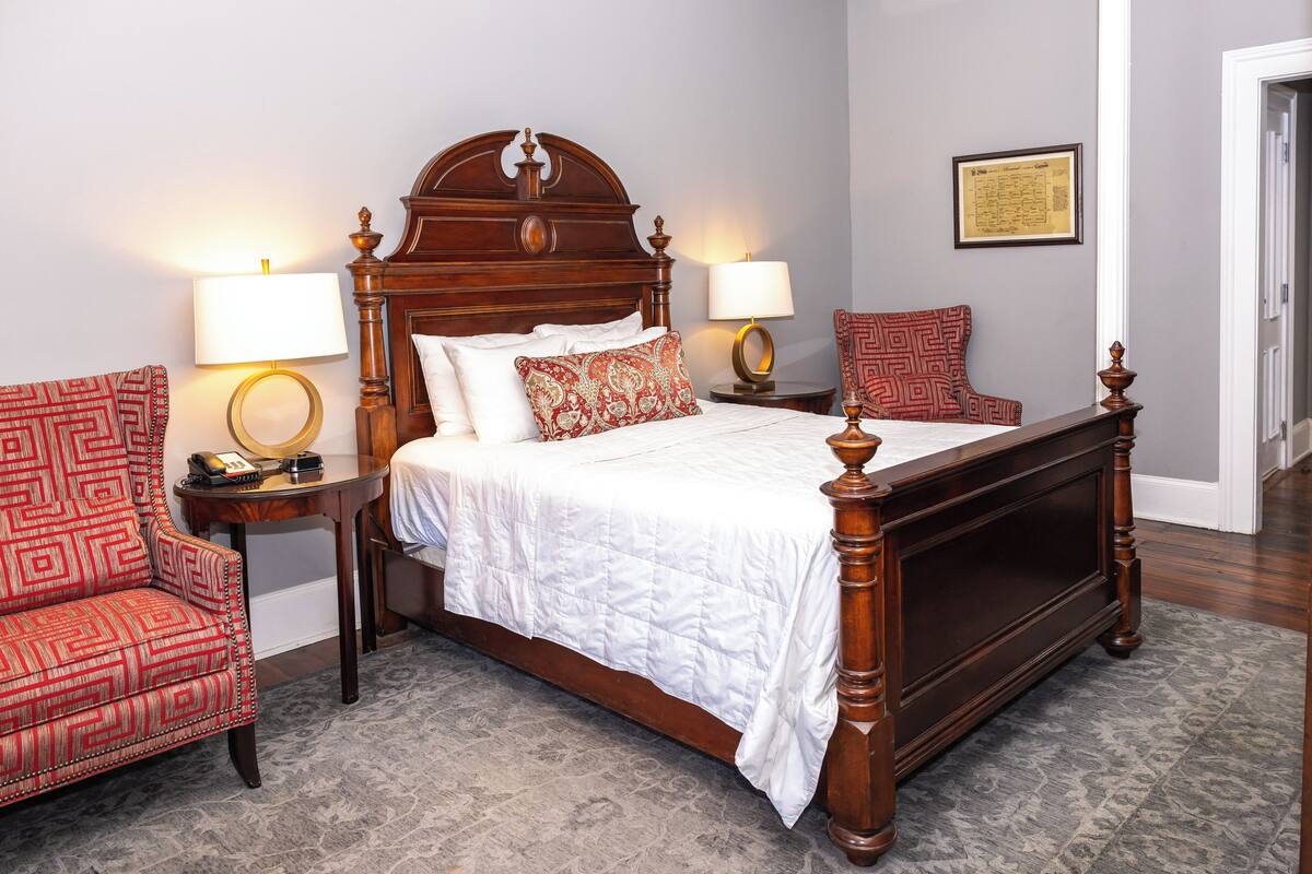 A neatly arranged bedroom with a large bed, two bedside tables with lamps, a patterned armchair, and a framed certificate on the wall.