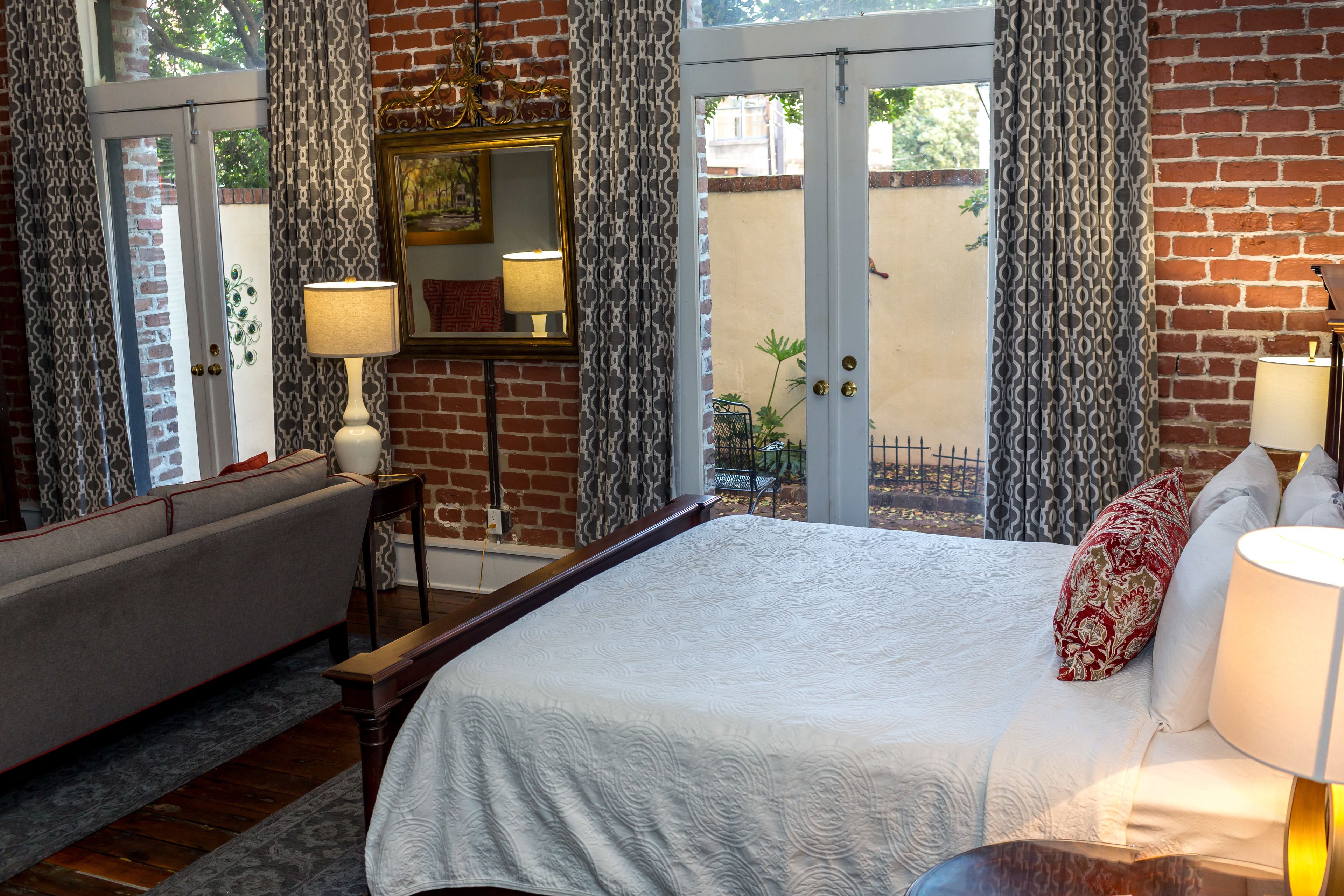 A bedroom with a large bed, a sitting area with a sofa, a table, and a chair. There is a window with curtains and a view of the outside.