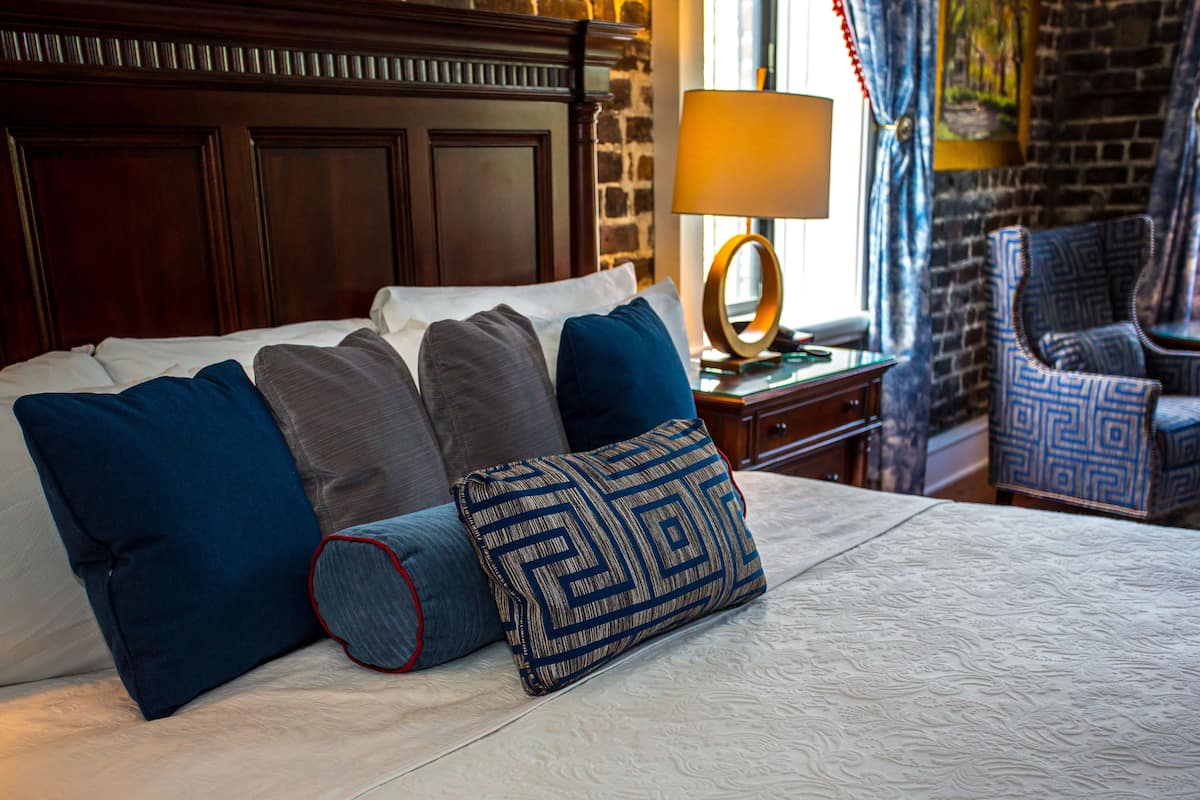 A neatly made bed with multiple pillows, a wooden headboard, a bedside table with a lamp, and a patterned armchair.